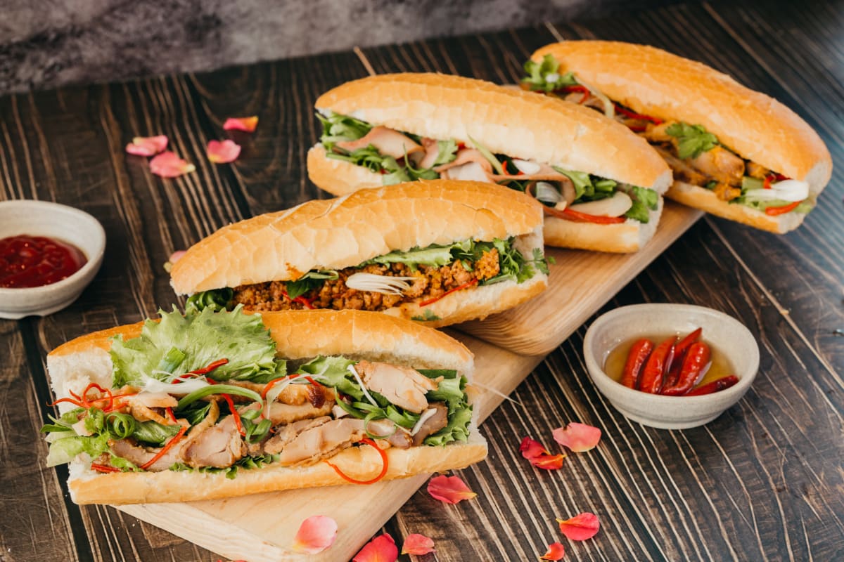 Four open rolls/mini baguettes, with Asian style ingredients, a variety of meats, and salad. Served on a wooden chopping board with pickled chillis and a red hot sauce 