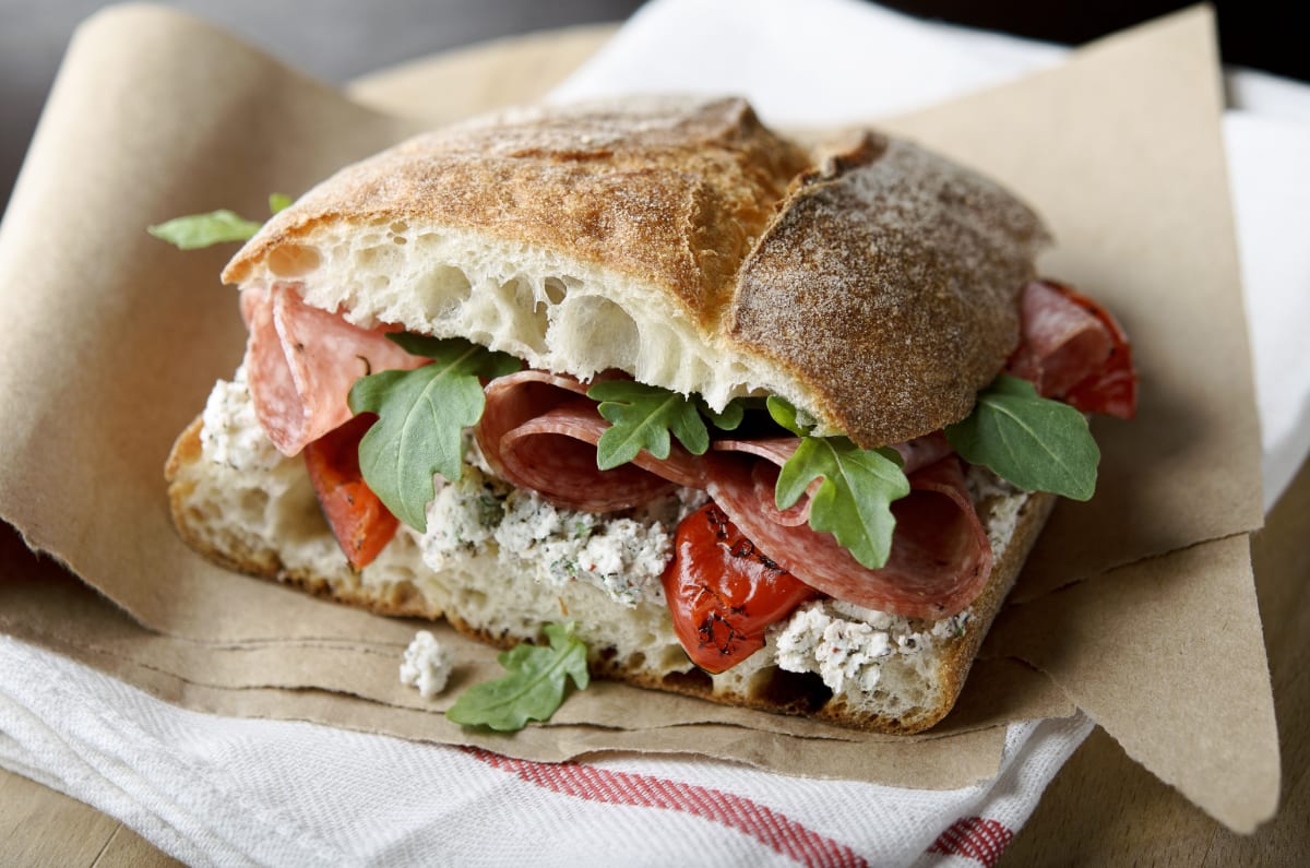 Traditional Italian sandwich, served on a focaccia style bread roll with arugula, tomatoes, and salami inside the bun. Served on brown paper, with a red and white tea towel below.  