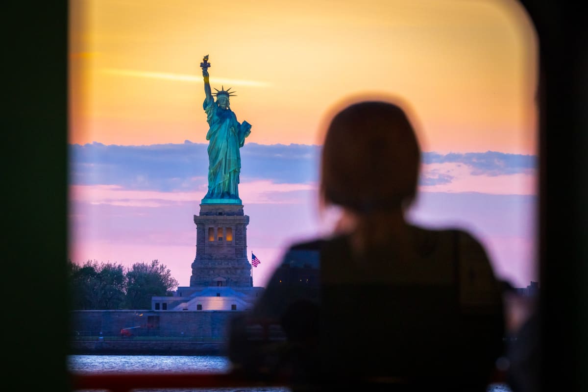 silhouette of person looking out of window towards statue of liberty at sunset 