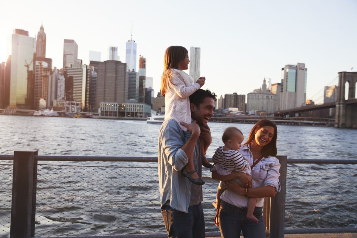 Family of mother, father, young girl and baby stand smiling in front of the waterfront and New York skyline