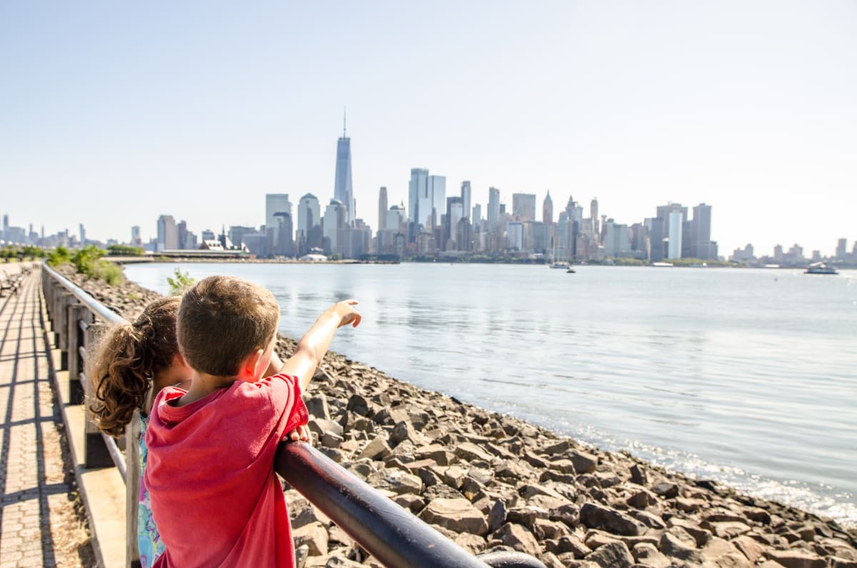 Young girl and boy stand with back to camera looking and pointing across the water to the New York skyline