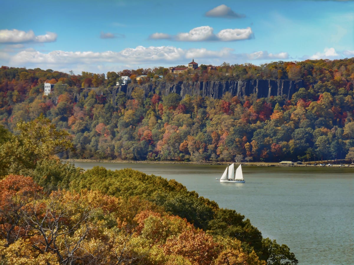 Sail boat sails on the Hudson river between two tree covered banks with fall colors