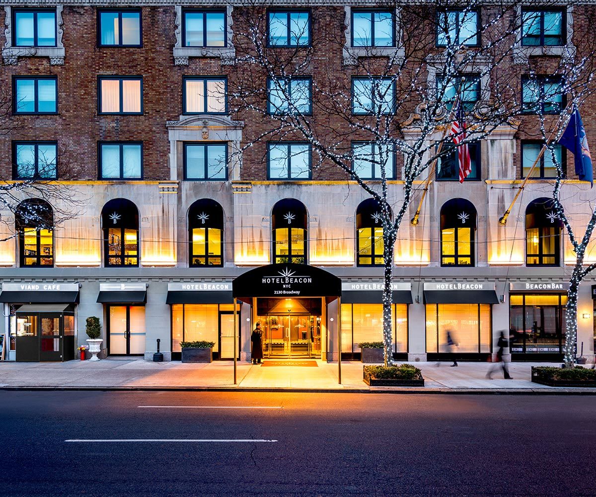 hotel beacon in new york, red brick and concrete building, with lights in the trees in front, american flag hangs from flag pole. porter stands at hotel entrance