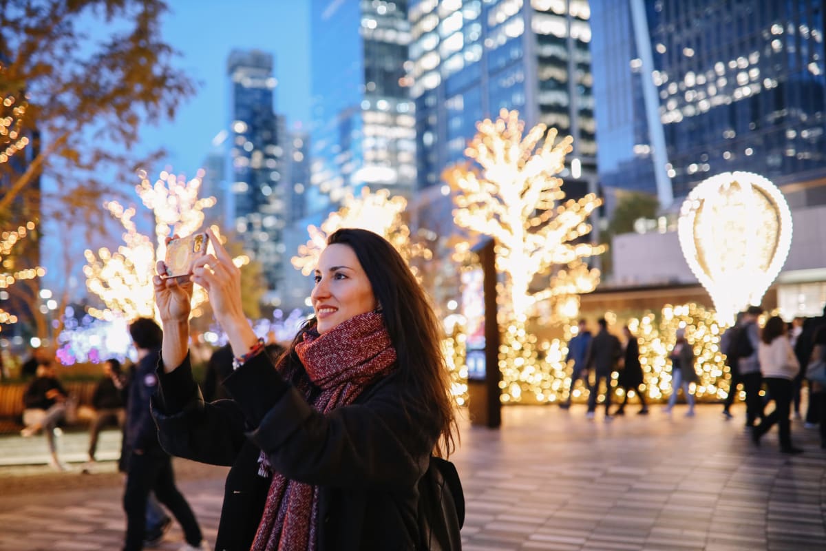  Woman takes a photo with her phone while surrounded by lit up trees, skyscrapers and other christmas decorations