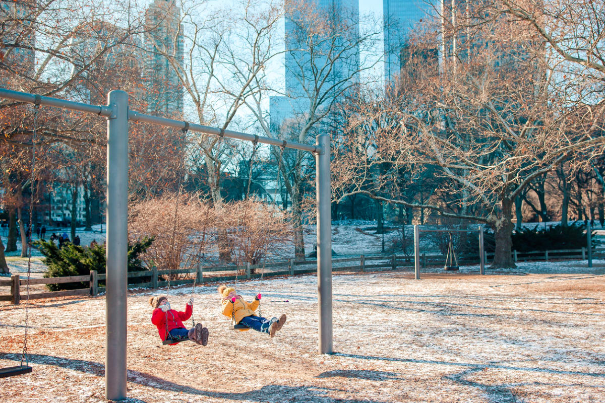 Kids in red and yellow jackets swing on the swings in a park in front of skyscrapers in the snow