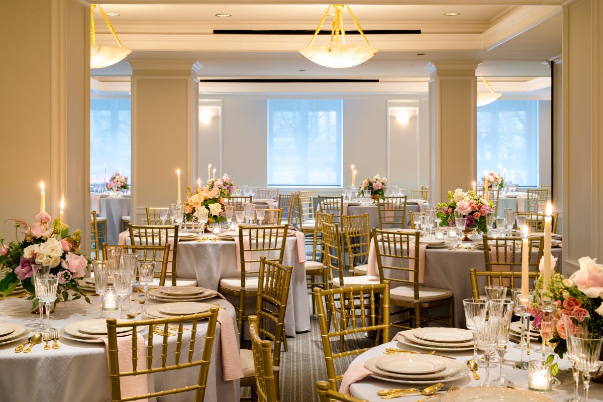 A large, elegantly set dining room with round tables, gold chairs, and flower centerpieces.