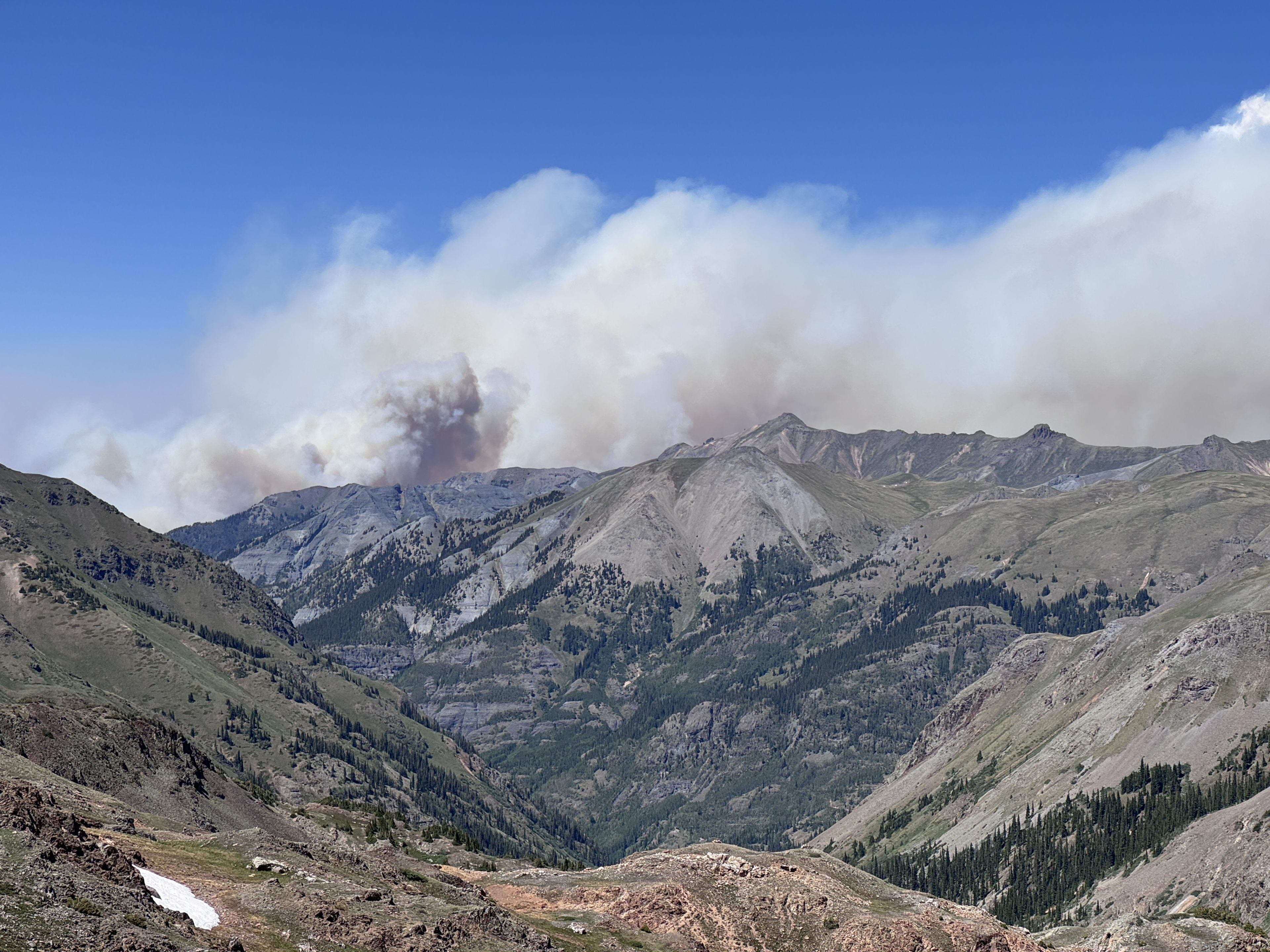 A large wildfire smoke plume rising behind a green ridge in daylight
