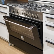 View of a contemporary kitchen which features a cabinetry, countertop, drawer, floor, flooring, furniture, gas stove, hardwood, home appliance, kitchen, kitchen appliance, kitchen stove, major appliance, brown, white
