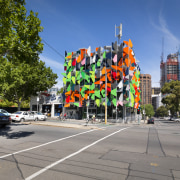 View of Pixel Building, with colorful exterior flags, car, city, downtown, landmark, metropolis, metropolitan area, mixed use, neighbourhood, pedestrian, road, sky, street, tourist attraction, town square, tree, urban area, gray