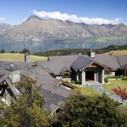 View of contemporary home by lake and mountains. alps, cottage, elevation, estate, hill station, home, house, landscape, mount scenery, mountain, mountain range, mountain village, mountainous landforms, real estate, residential area, roof, rural area, sky, suburb, tree, village, wilderness, gray