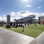 Paved path and grass area with people. - architecture, building, city, daytime, downtown, grass, metropolitan area, mixed use, recreation, sky, structure, town square, urban area, urban design, teal