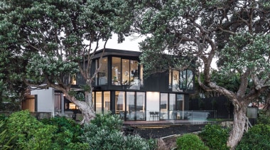This new home in Whangaparaoa is nestled amongst architecture, cottage, estate, home, house, outdoor structure, plant, property, real estate, residential area, tree, villa, black