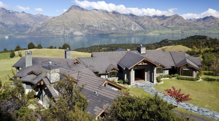 View of contemporary home by lake and mountains. alps, cottage, elevation, estate, hill station, home, house, landscape, mount scenery, mountain, mountain range, mountain village, mountainous landforms, real estate, residential area, roof, rural area, sky, suburb, tree, village, wilderness, gray