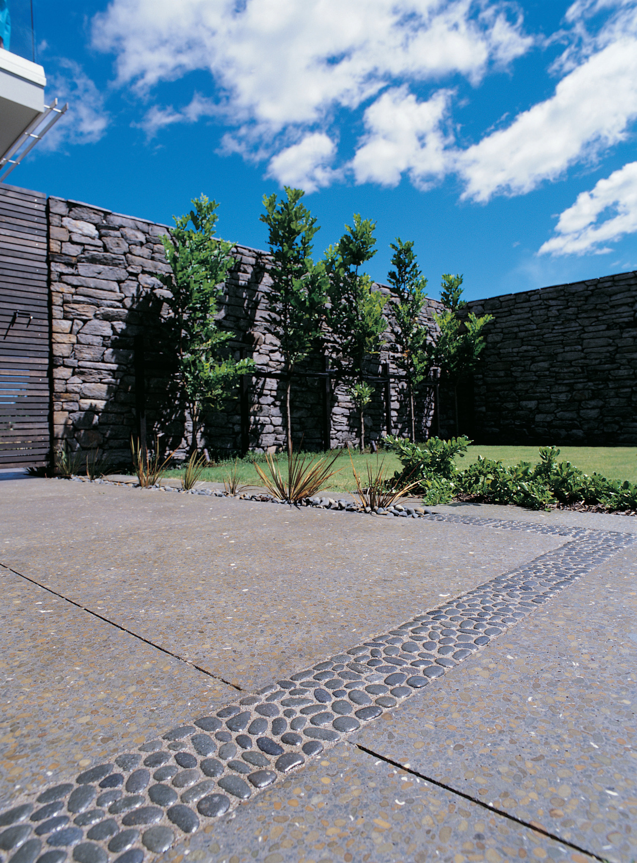 Paved area with exposed aggregate concrete and contrasting architecture, cobblestone, courtyard, driveway, estate, facade, grass, home, house, landscape, landscaping, outdoor structure, plant, real estate, residential area, road surface, sky, stone wall, tree, walkway, wall, gray