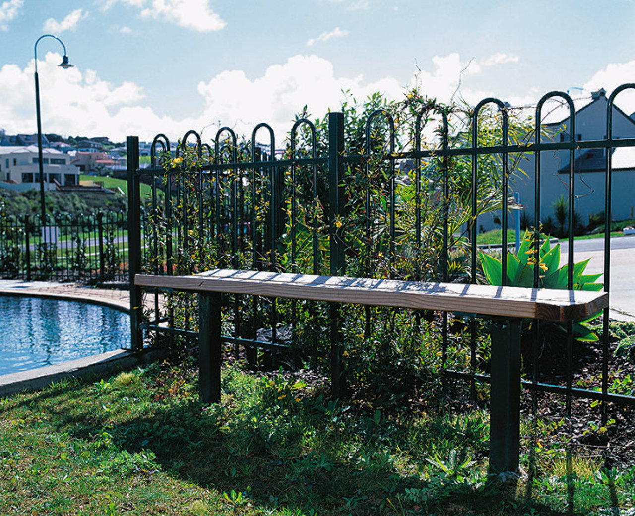 Bench seat in front of steel fence. fence, grass, outdoor structure, plant, real estate, tree, water, white, black