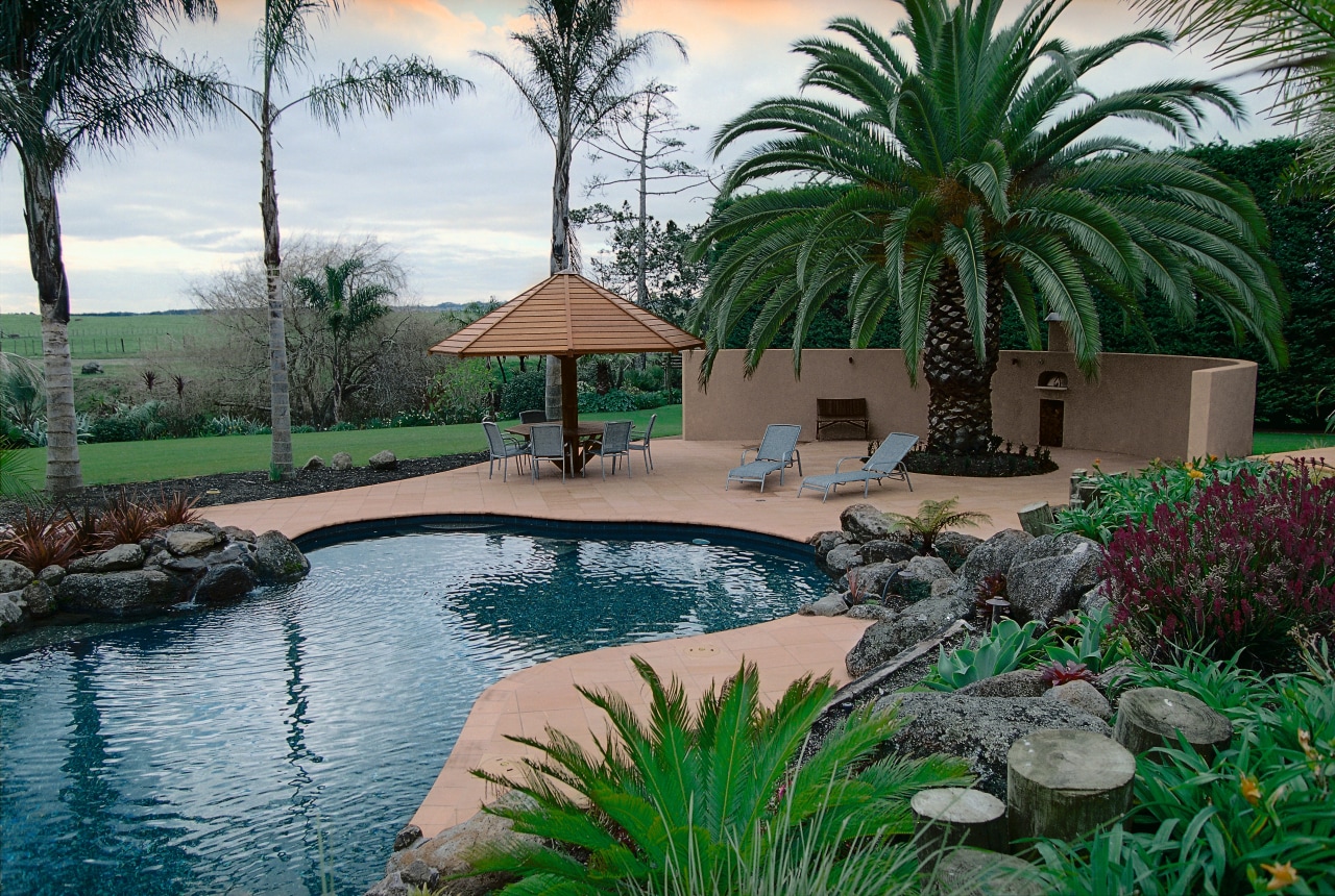 view of this lush outdoor living/pool area featuring arecales, backyard, estate, hacienda, home, landscape, landscaping, leisure, outdoor structure, palm tree, plant, property, real estate, resort, swimming pool, tree, water feature, yard, gray
