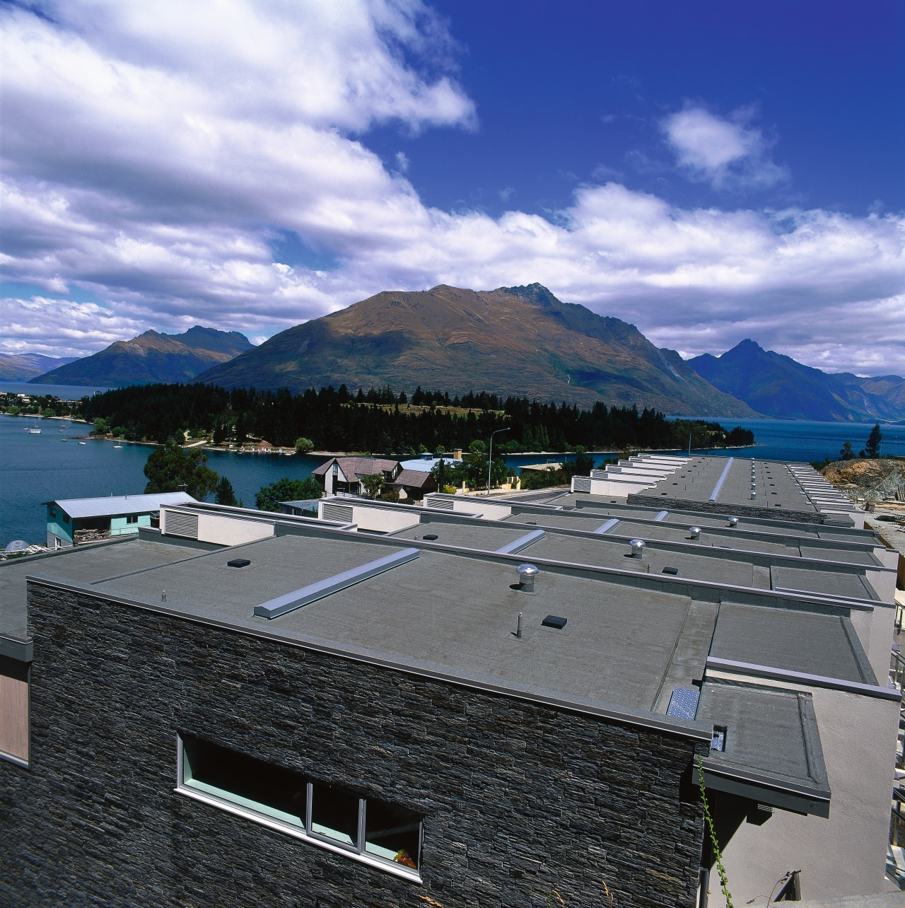 View of apartment building grey roofing with stone architecture, cloud, house, lake, mountain, mountain range, outdoor structure, real estate, roof, sea, sky, water, blue, black