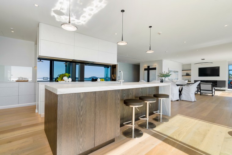The mirrored splashback behind the cooktop reflects the architecture, building, cabinetry, ceiling, countertop, design, dining room, floor, flooring, furniture, hardwood, home, house, interior design, kitchen, mirror splashback,  mirror backsplash, wood flooring, property, real estate, room, table, wood, wood flooring, gray, white