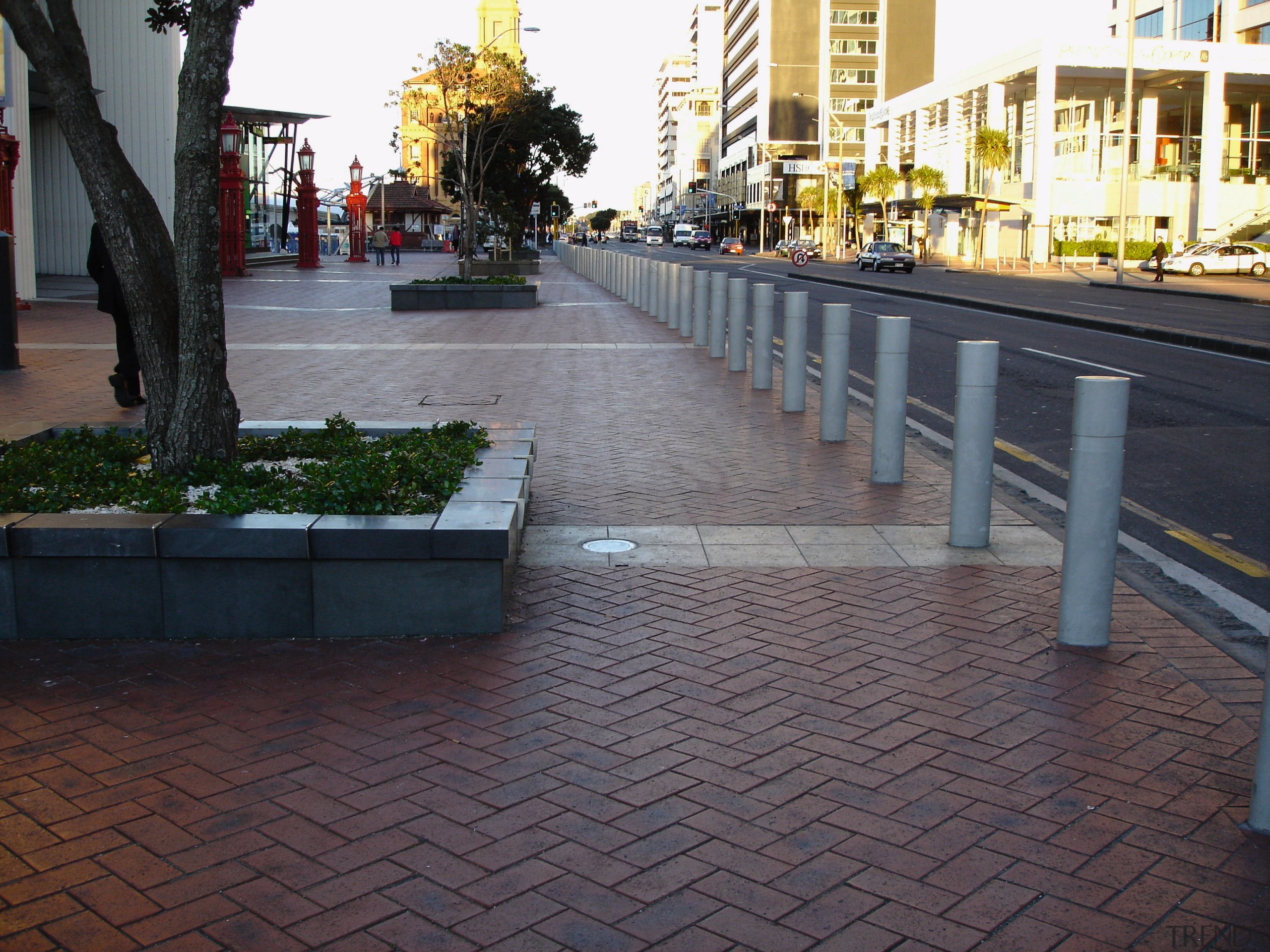 Patterned paved footpath with bollards, and raised gardens. boardwalk, cobblestone, floor, flooring, lane, neighbourhood, outdoor structure, public space, road surface, sidewalk, tree, urban design, walkway, wall, gray, black