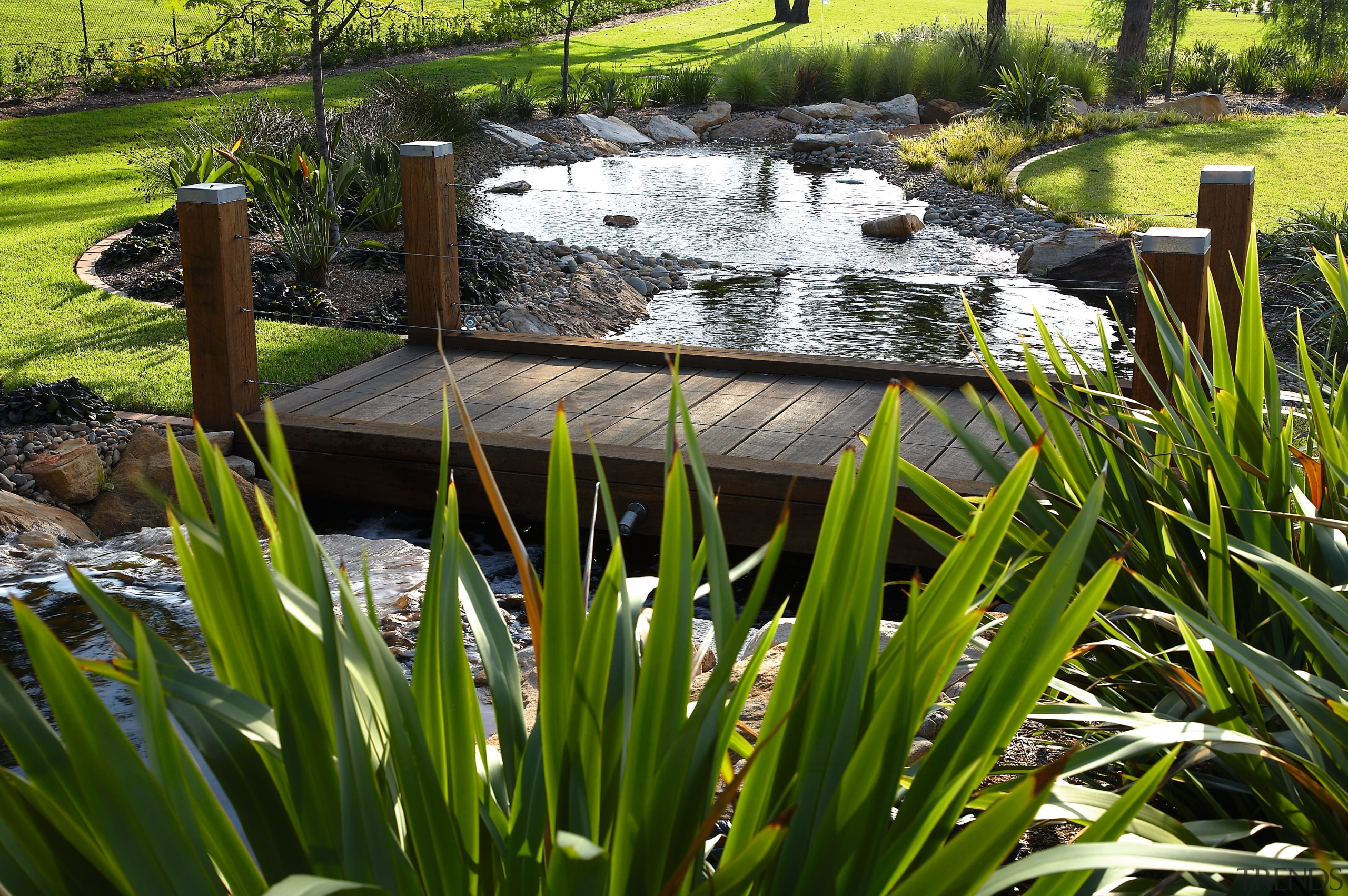 View of this timber boardwalk bridge crossing the backyard, flower, garden, grass, landscape, landscaping, outdoor structure, plant, pond, water, water feature, brown