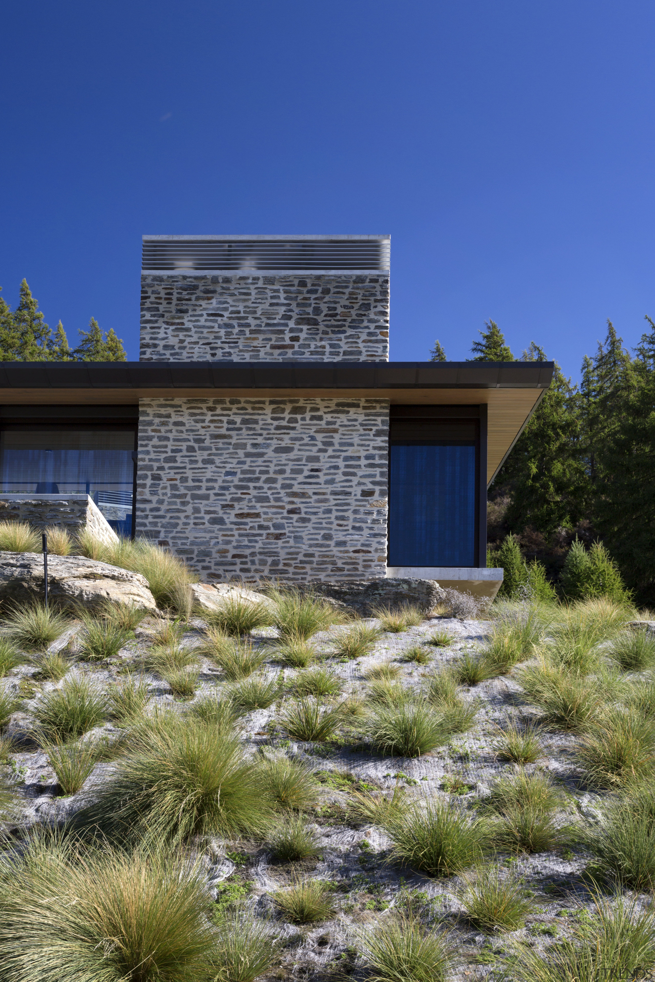 On this alpine home, the material palette has architecture, cottage, elevation, estate, facade, grass, grass family, home, house, land lot, landscape, landscaping, property, real estate, residential area, sky, blue, brown