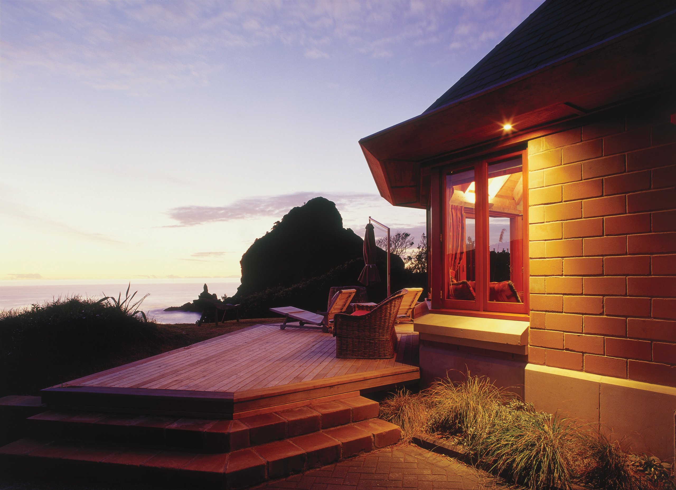 An exterior view of this coastal piha home architecture, cottage, dusk, estate, evening, home, house, lighting, morning, property, real estate, roof, sky, sunlight, sunset, window, wood, red