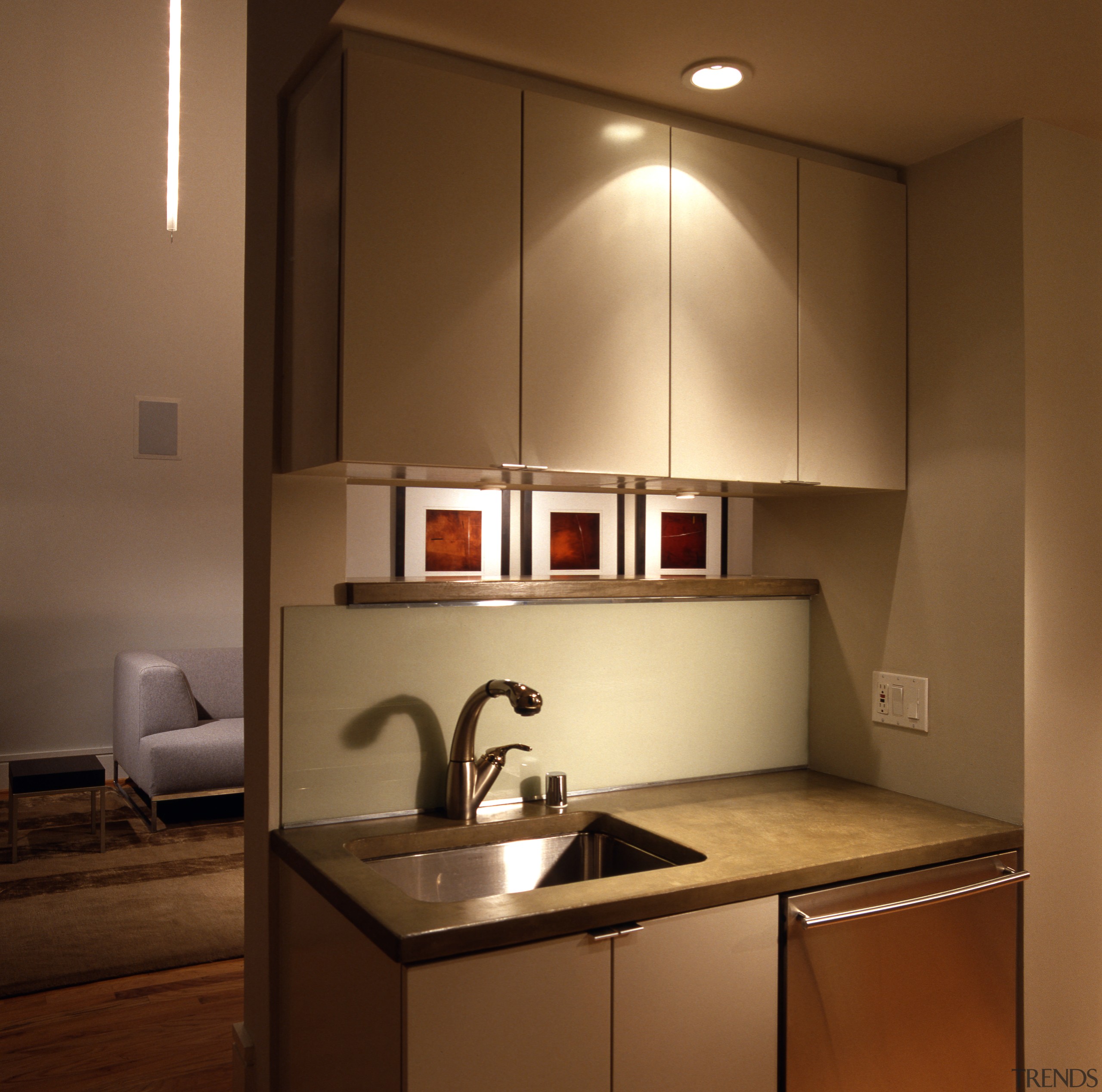 view of kitchen and living area, featuring cabinetry, cabinetry, ceiling, countertop, interior design, kitchen, light fixture, lighting, room, under cabinet lighting, brown