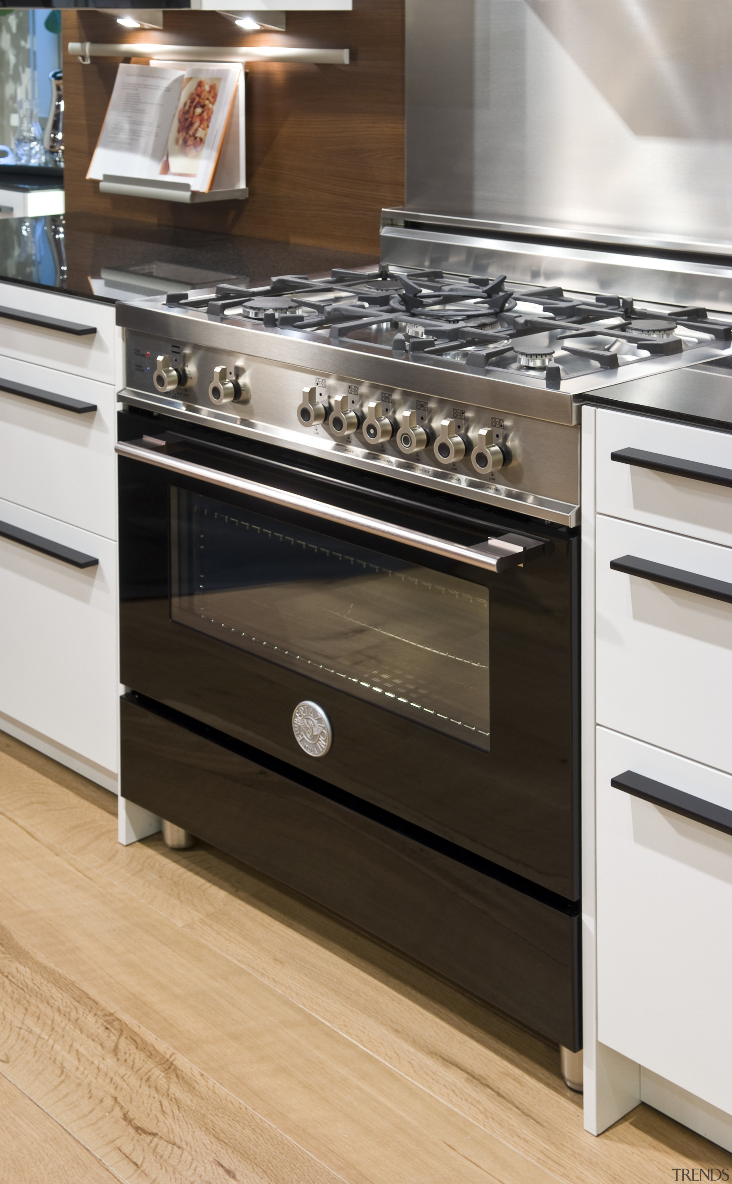 View of a contemporary kitchen which features a cabinetry, countertop, drawer, floor, flooring, furniture, gas stove, hardwood, home appliance, kitchen, kitchen appliance, kitchen stove, major appliance, oven, brown, white