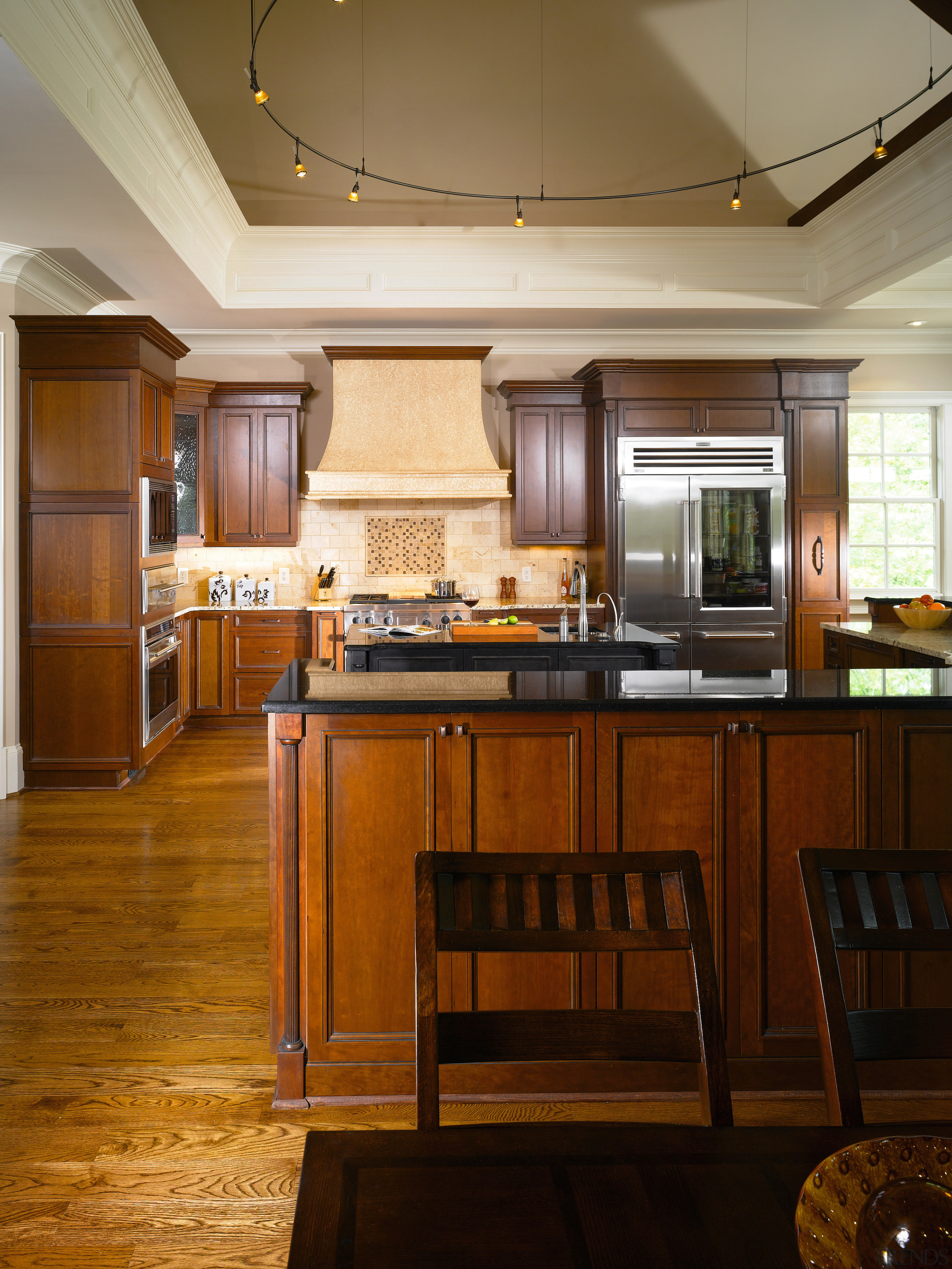 The oversized range hood connects tonally with adjacent cabinetry, ceiling, countertop, cuisine classique, floor, flooring, hardwood, interior design, kitchen, room, wood, wood flooring, brown