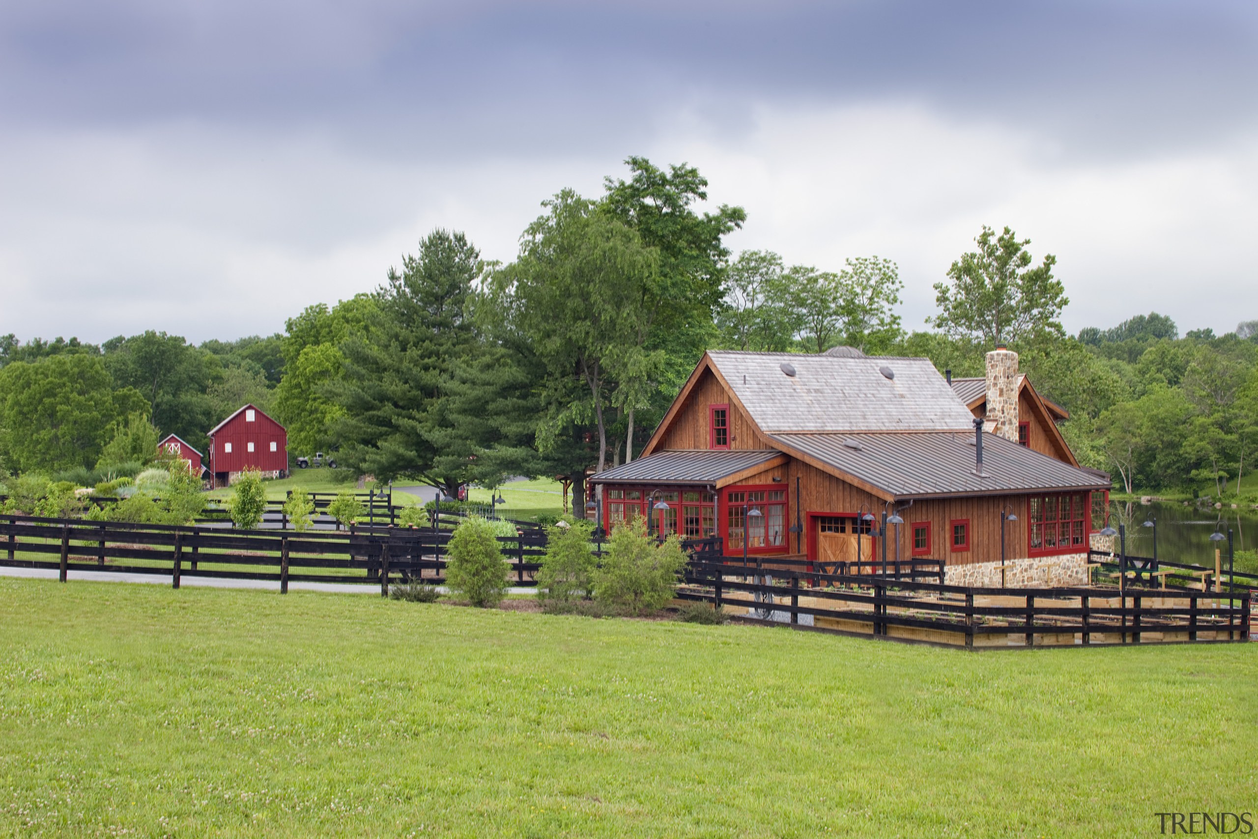 View of wooden building with black fence and barn, cottage, estate, farm, farmhouse, fence, field, grass, grassland, home, house, landscape, log cabin, meadow, pasture, ranch, real estate, rural area, sky, tree, green, gray