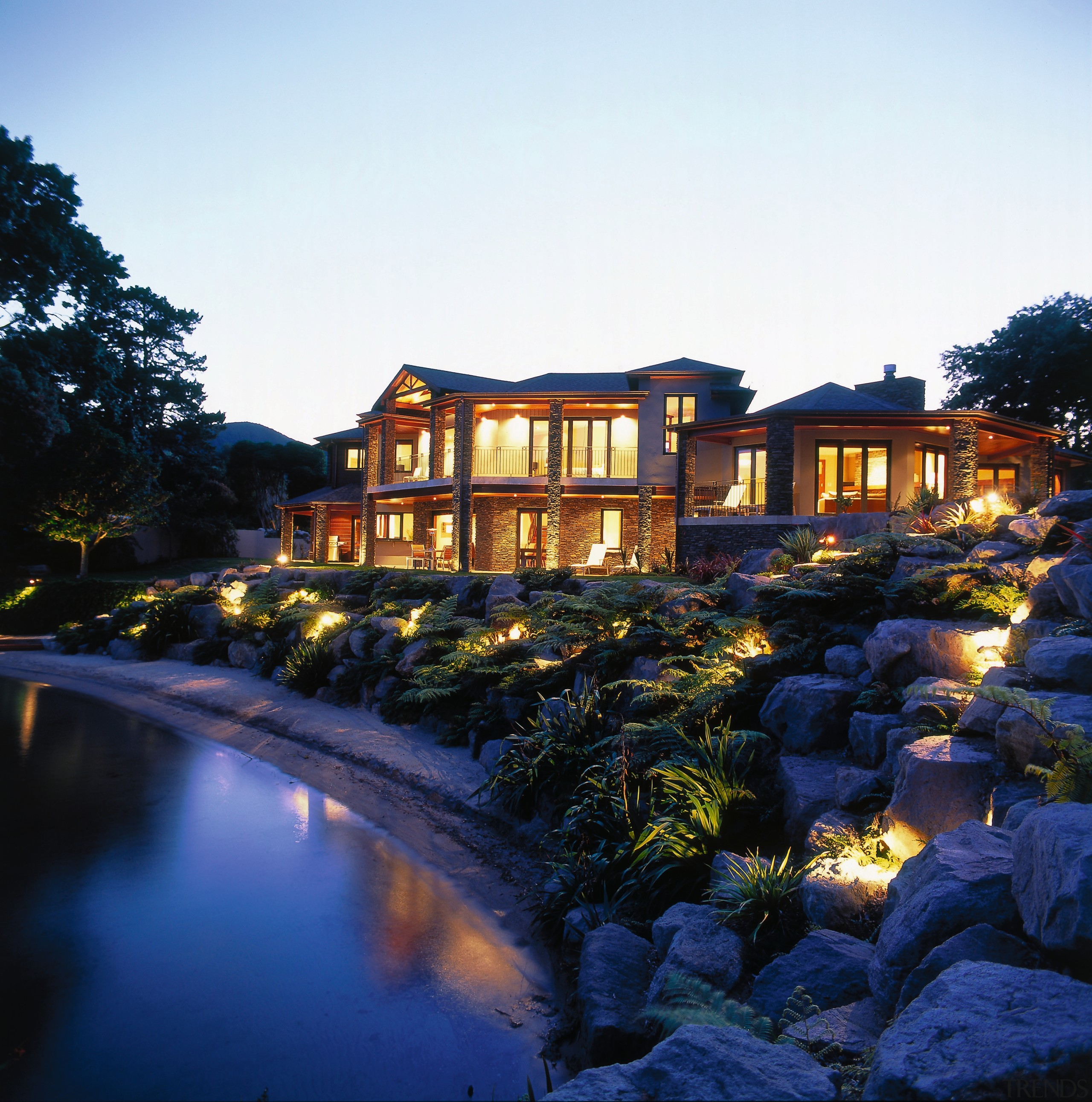 Side on view of lakeside property. Features rock architecture, estate, evening, home, house, landscape, landscape lighting, lighting, mansion, morning, night, property, real estate, reflection, residential area, resort, sky, tree, villa, water, white, black