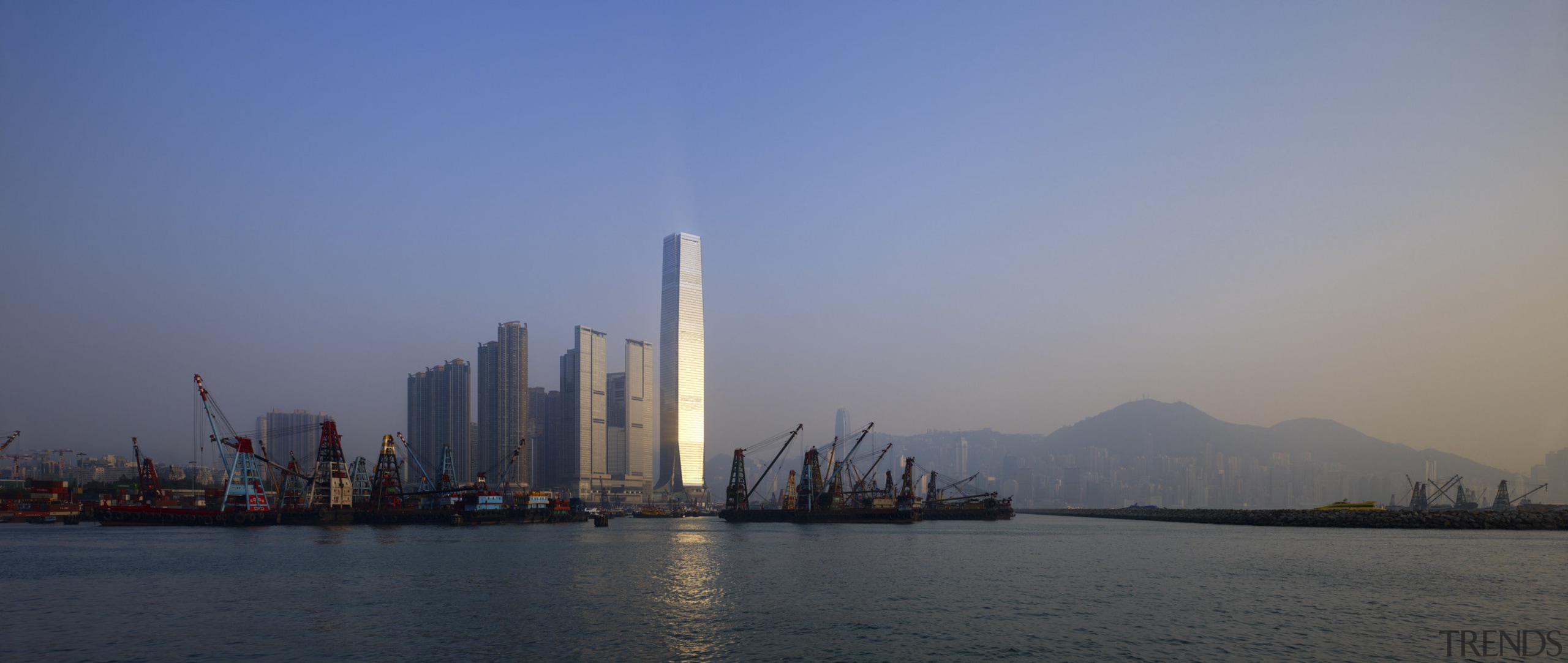 Here is a view of the ICC (International body of water, calm, city, cityscape, dawn, daytime, dusk, evening, haze, horizon, metropolis, metropolitan area, morning, reflection, river, sea, sky, skyline, skyscraper, sunset, tower, tower block, water, blue, gray