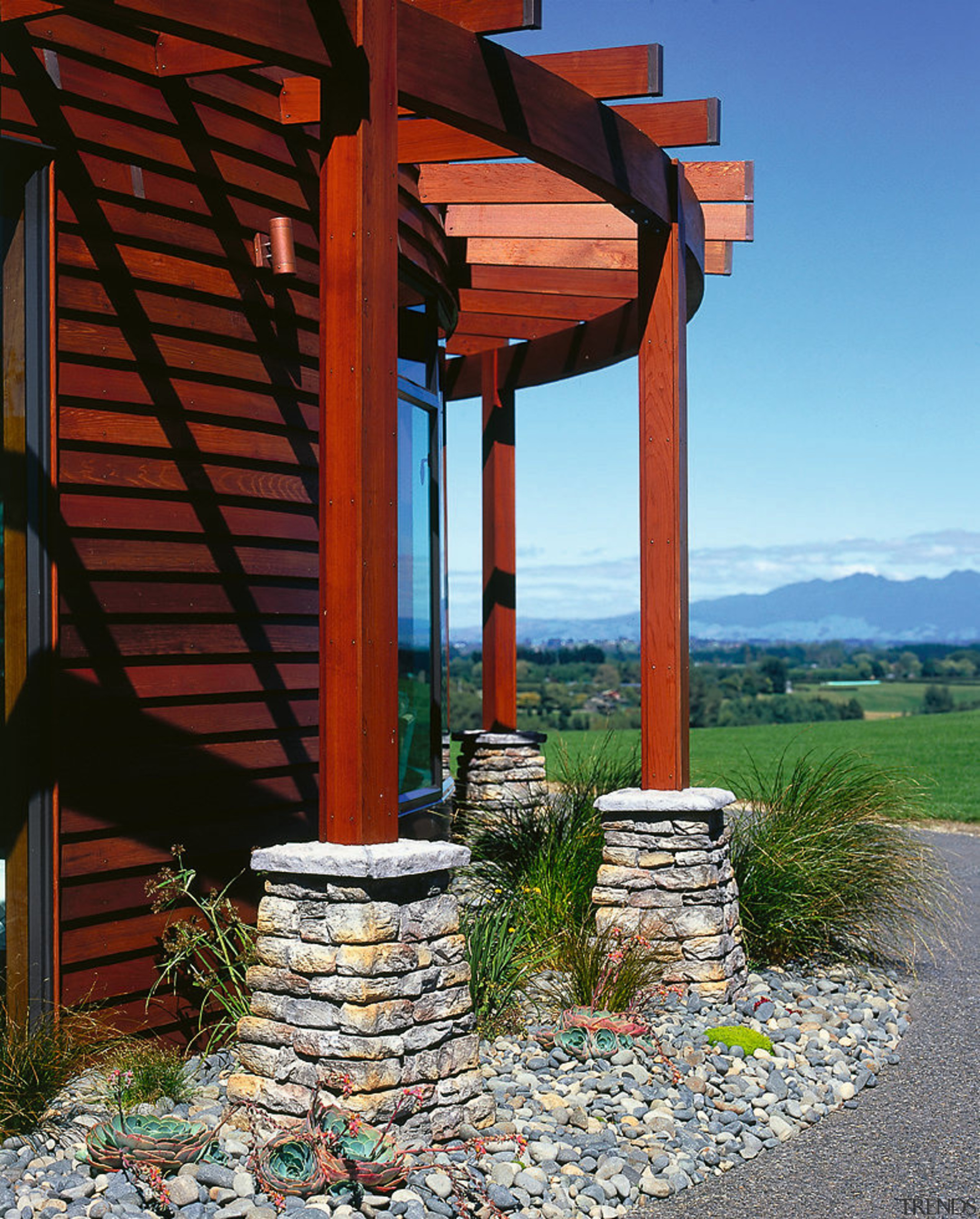 A photograph of kiwi schist columns. There is house, outdoor structure, sky, walkway, wood, red