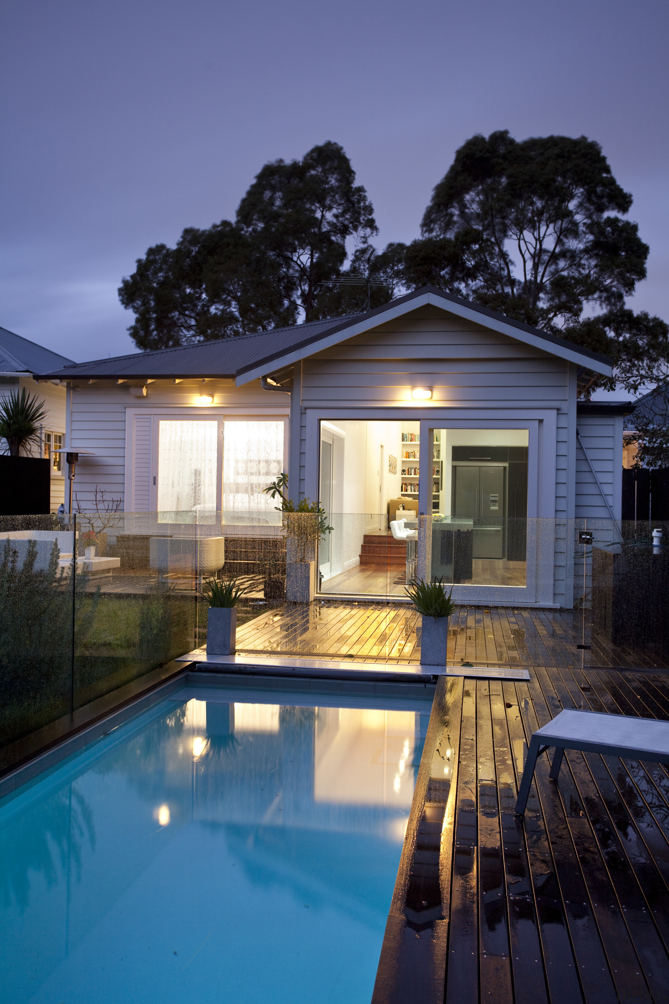 View of contemporary home with wooden flooring, glass architecture, backyard, cottage, estate, evening, facade, home, house, landscape lighting, lighting, property, real estate, reflection, residential area, siding, sky, swimming pool, villa, window, blue