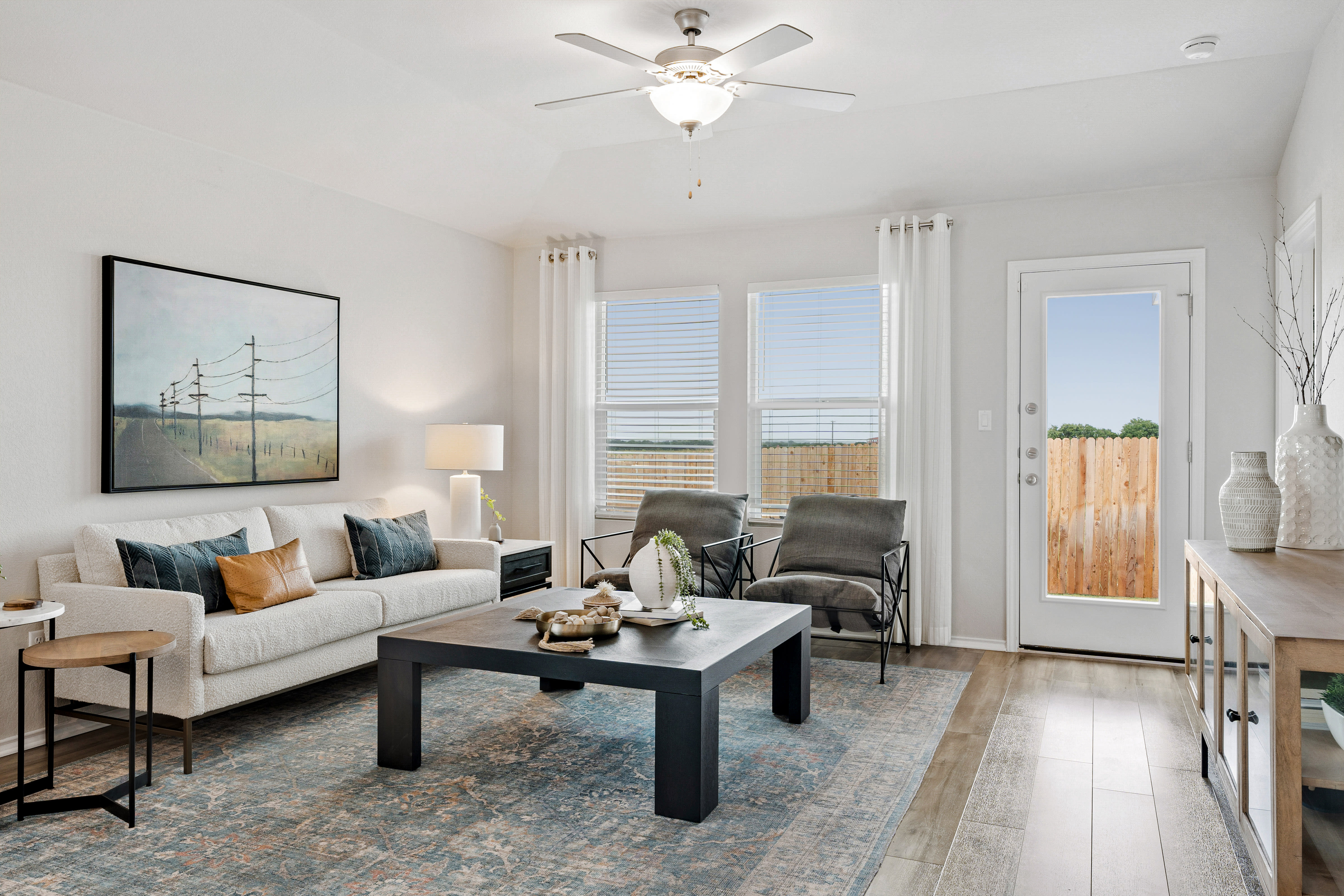 Photo of staged living room featuring ceiling fan, wood-style flooring, two-inch wood-style blinds and view of backyard fence.