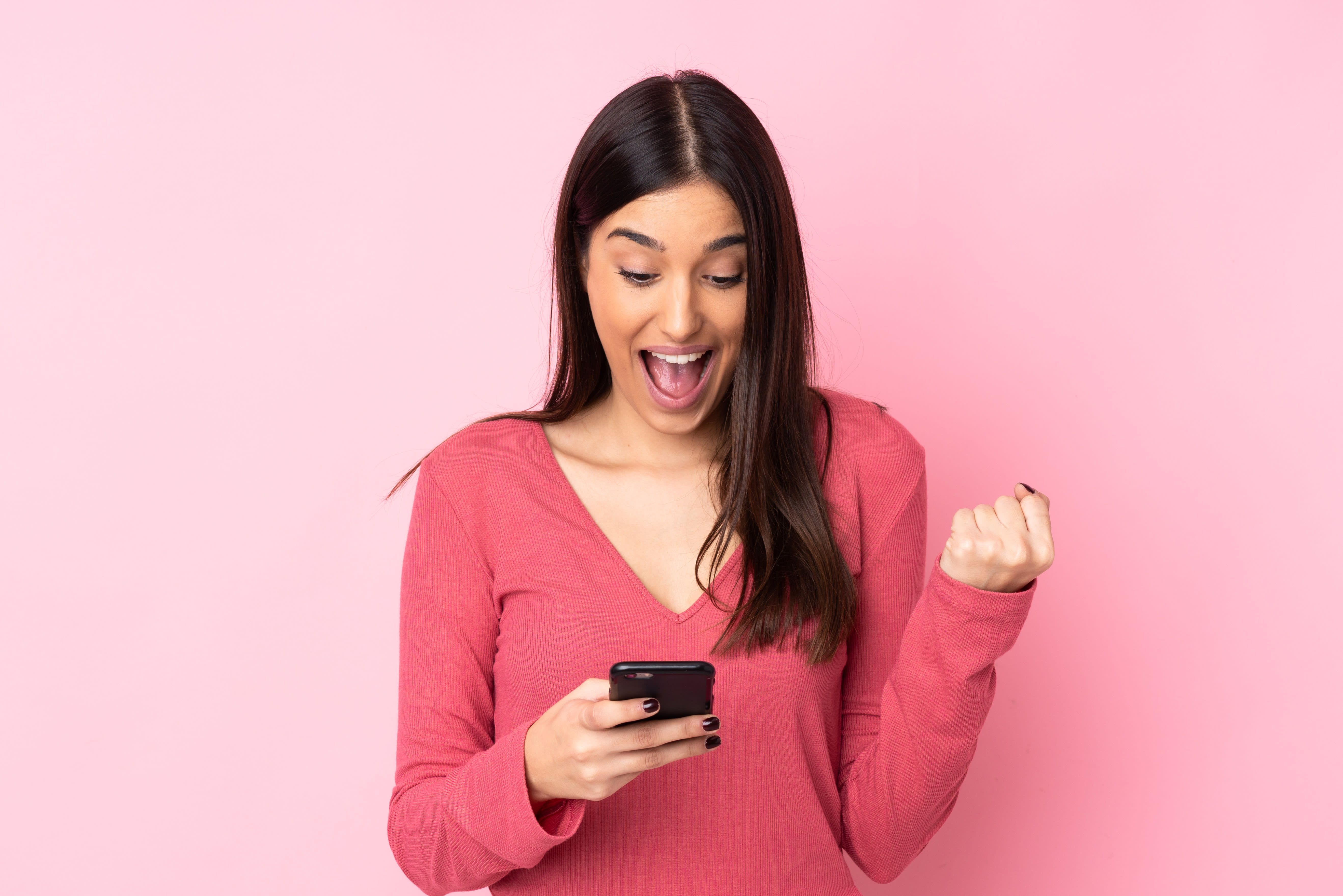 Woman looking at her phone excitedly against pink background