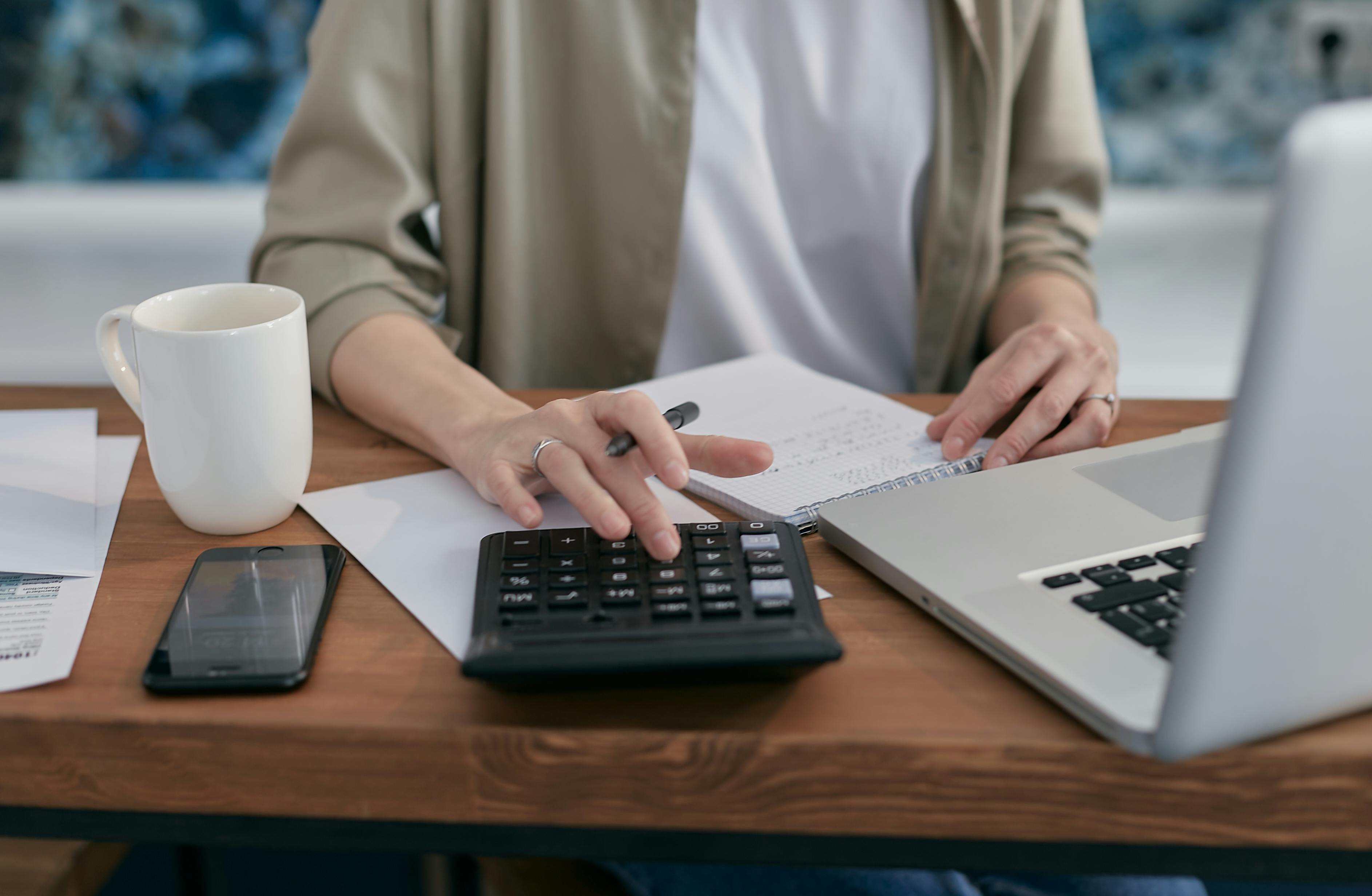 Frau am Schreibtisch mit Taschenrechner, Handy, Laptop und Kaffee, die den Verpflegungsmehraufwand 2024 berechnet