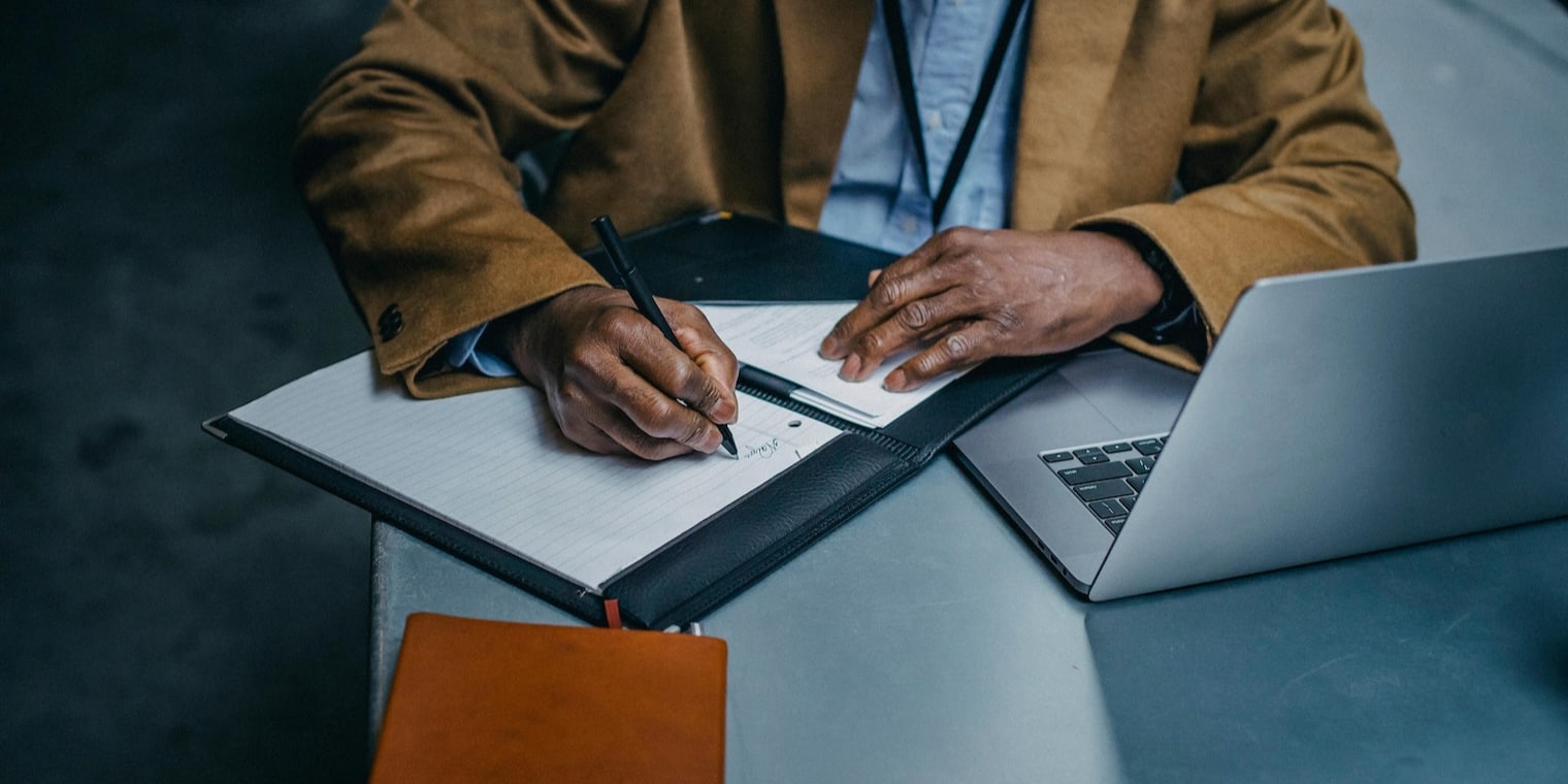 Business travel expenses list. Hands writing on white paper next to laptop.
