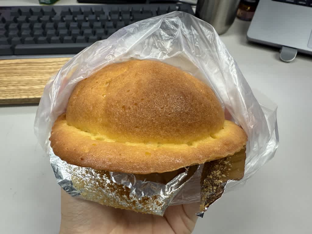 手に持った菓子パンがビニール袋に入っている。焼き色が均一でふっくらした丸い帽子のような形のパンで、底面にアルミホイルが敷かれている