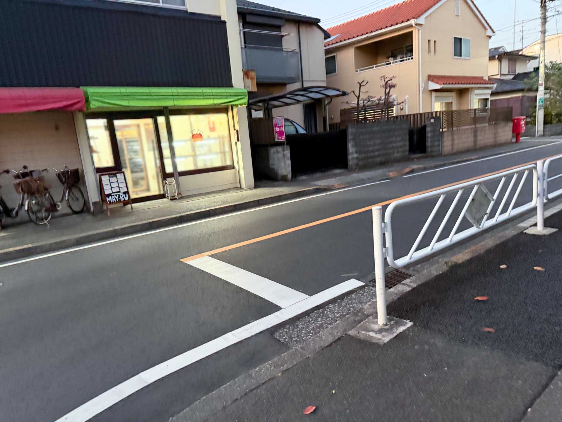 目的のパン屋の外観。住宅街の道路を挟んで向こう側にある。緑と赤のひさしの下にガラス張りの入り口があり、店舗前には小さな看板が置かれている。左側には自転車が数台停められており、右側には住宅が並んでいる。