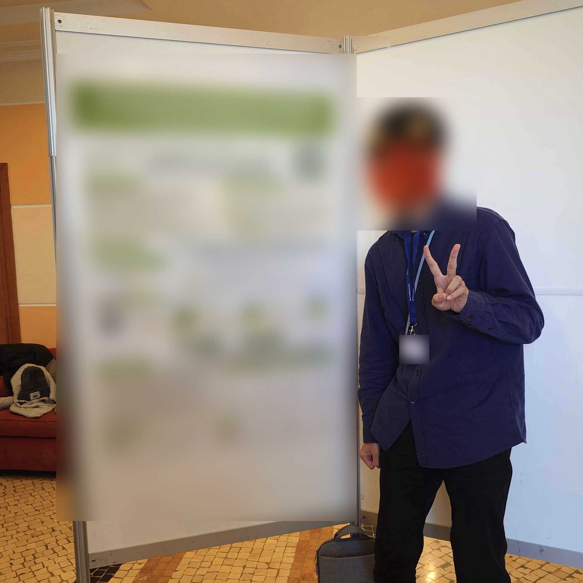 屋内の展示会場で、名札を下げた男性が仕切りパネルのポスター横に立ち、ピースサインをしている