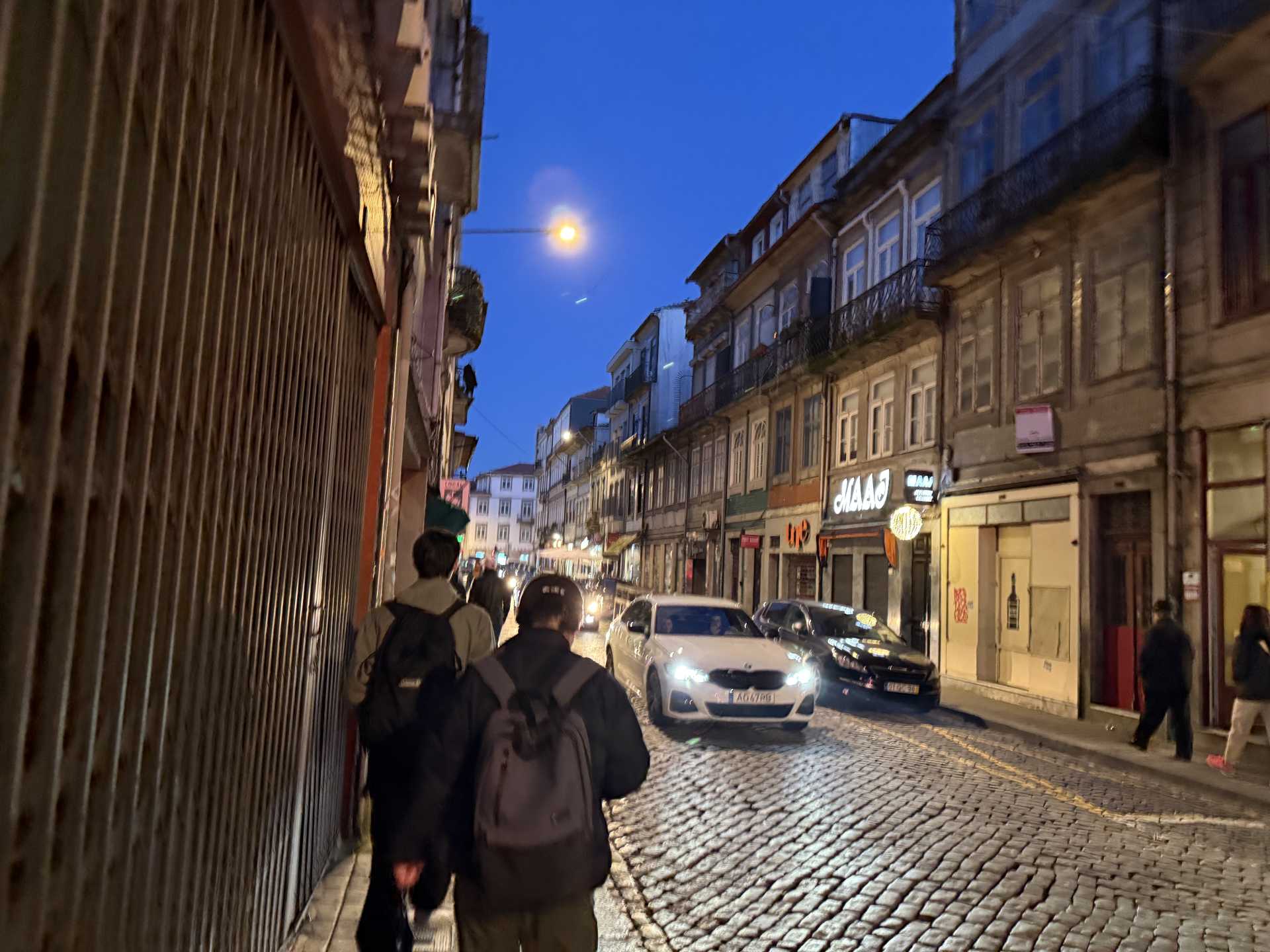 青い夕空の下、石畳の狭い通りに古い建物と店の看板が並び、車のヘッドライトが光る中、リュックを背負った人々が歩く。