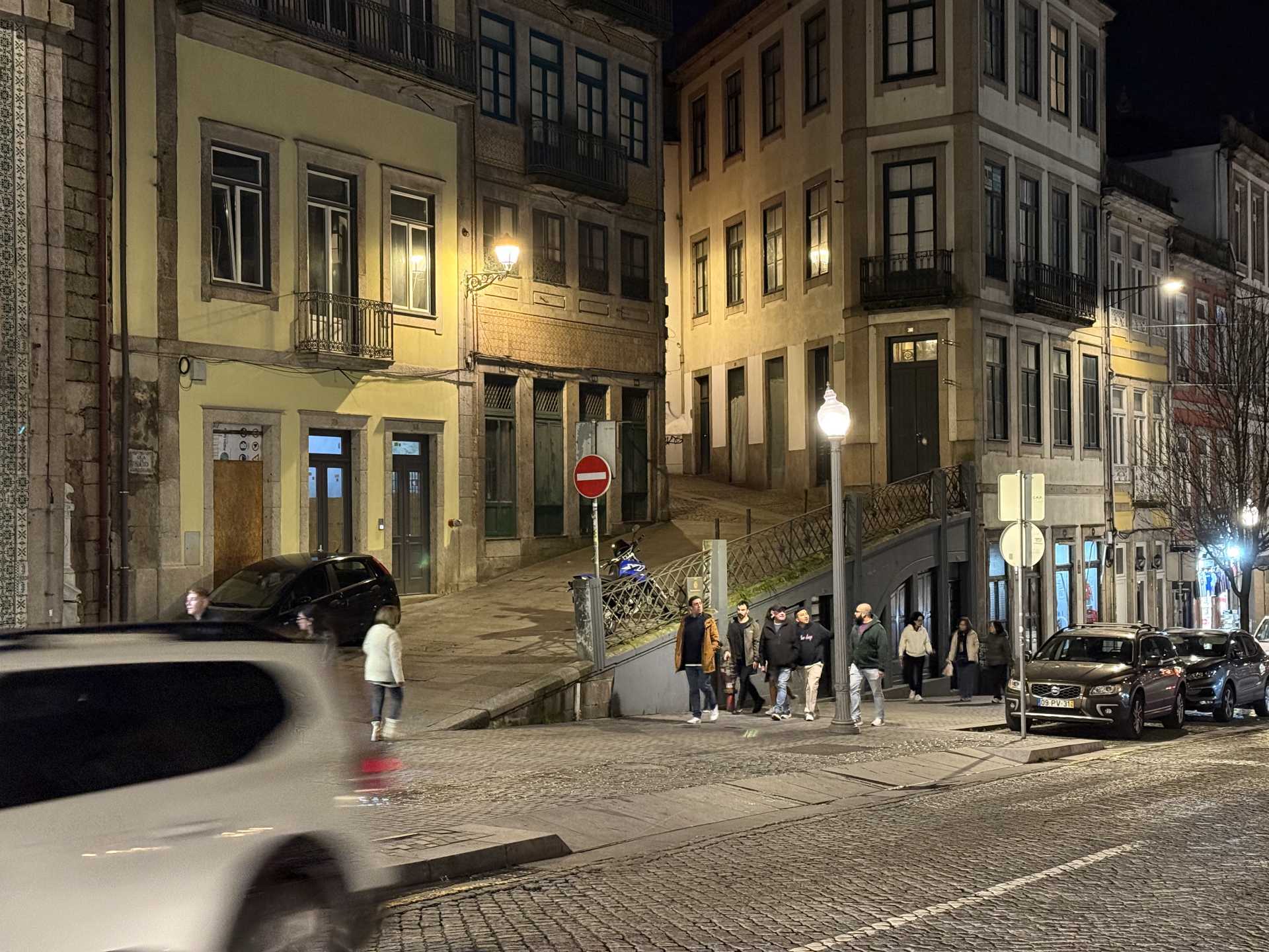 夜の石畳の坂道に古い建物が並び、街灯の下を歩行者が行き交い車が通る街角