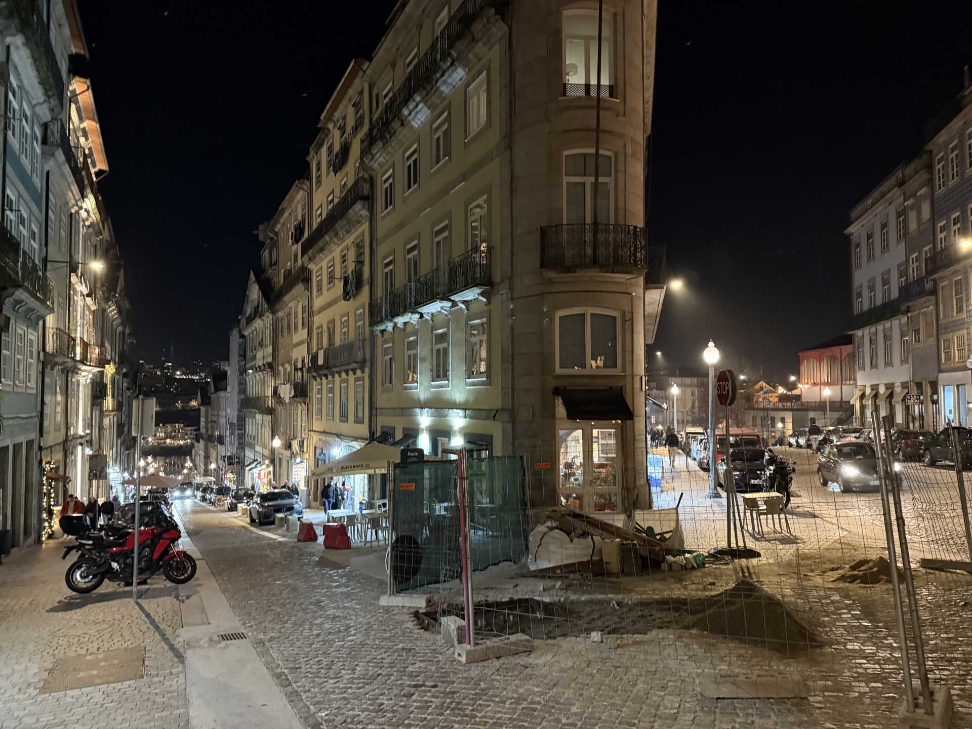 夜の石畳の坂道に古い建物が並び、街灯と車の灯りが照らす。角には工事用フェンスも見える。