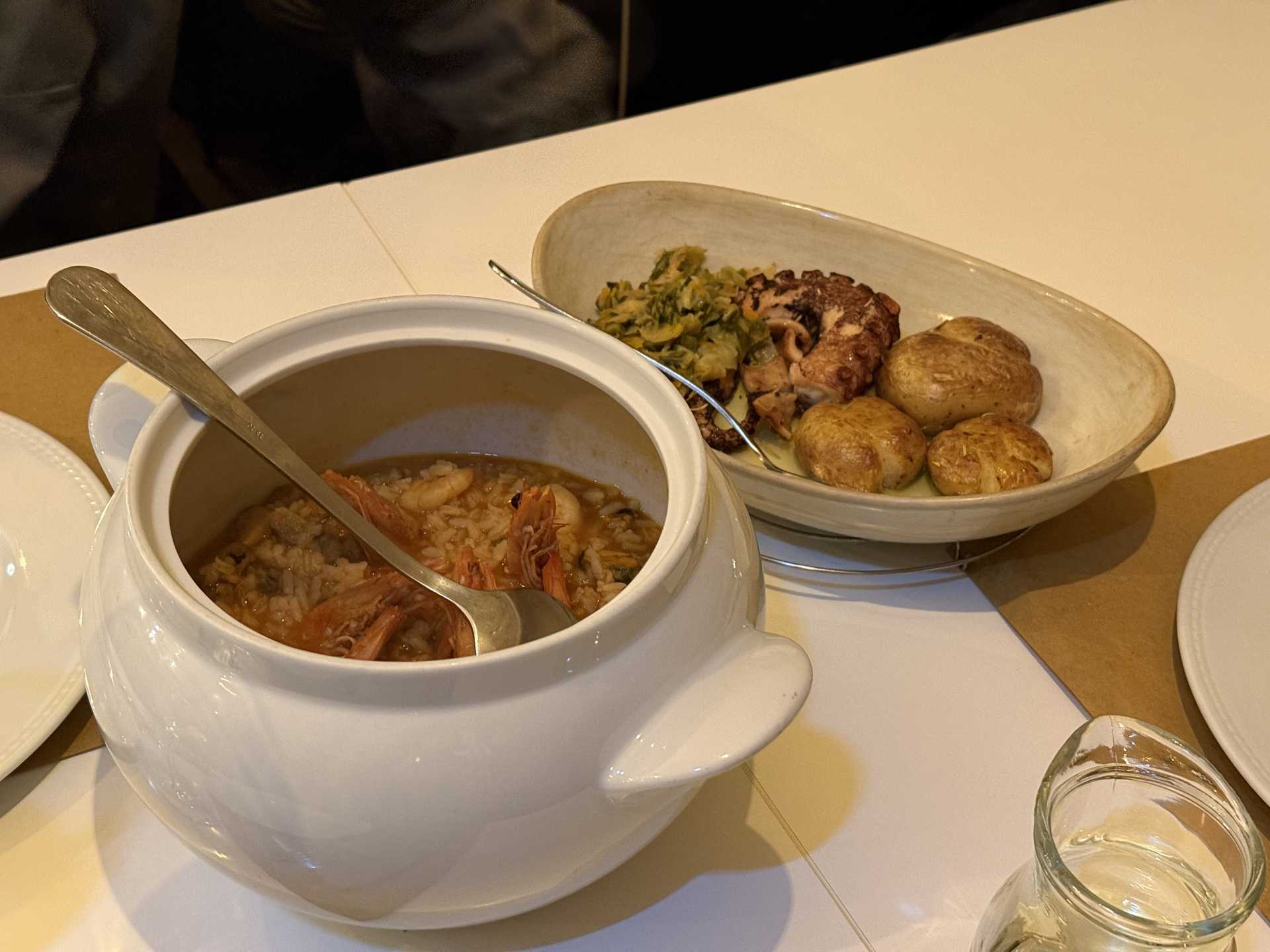 白い陶器鍋にエビ入りのスープご飯。横にタコのソテーと小じゃがいも、野菜の皿が並ぶ食卓。スプーンが差し込まれ、白ワインのグラスも見える。
