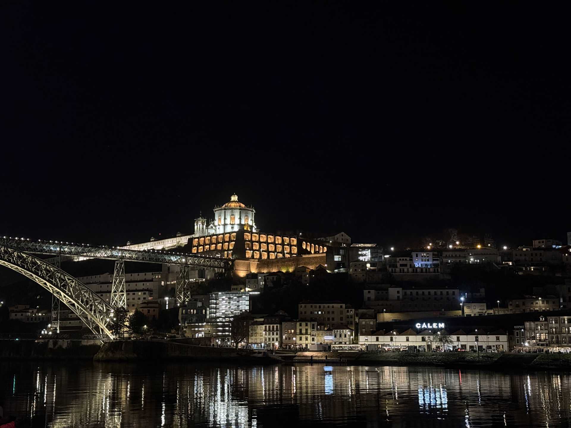 夜の川沿いの街並み。暗い空の下、ライトアップされた丘上の教会と鉄橋、岸辺の建物群の灯りが水面に反射して輝く静かな夜景。