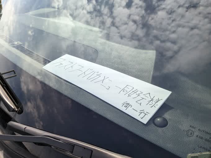 車のフロントガラスの部分に「ファストフードバカメニュー同好会様御一行」と書かれた紙が置かれている