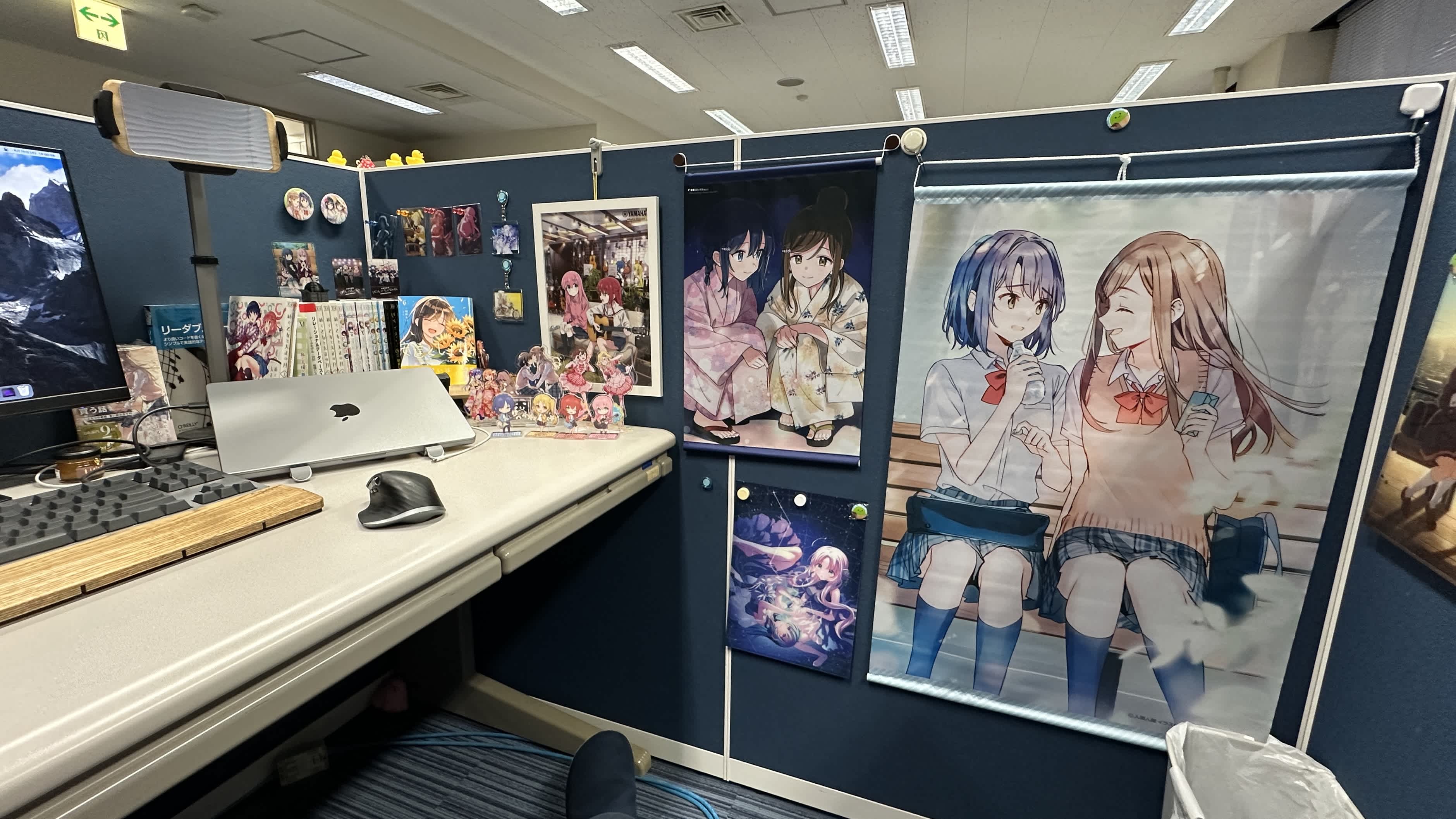 研究室の一角にあるデスク周りの写真。左手前には木製リストレスト付きのキーボード (Realforce) とワイヤレスマウス、そしてシルバーのノートPCスタンドに載った MacBook Pro が閉じて置かれている。奥のパーティションボードには濃い青色の仕切りがあり、そこにアニメや漫画のグッズが多数飾られている。具体的には、コミックス数冊が積まれ、その周囲にアニメキャラクターのアクリルスタンドフィギュア、缶バッジ、イラストカードなどが並んでいる。さらに、壁には浴衣姿の二人の少女を描いた『安達としまむら』のタペストリーがかけられている。右下にゴミ箱が置かれている。机の下の奥には、ピンク色のメンダコのぬいぐるみがひっそりと置かれている。全体としてアニメ関連の装飾に囲まれたデスクスペースが映し出されている。