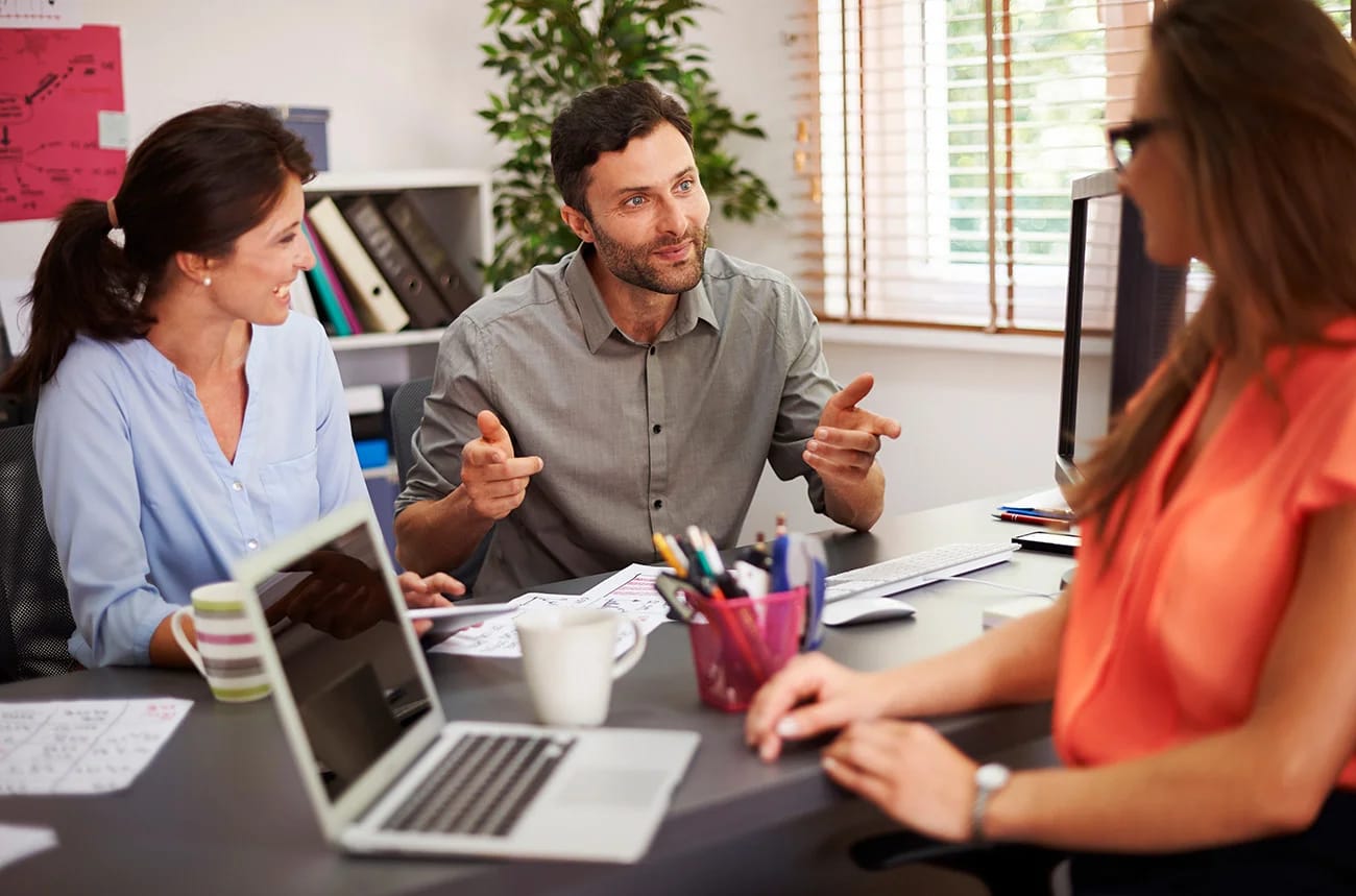 A small team talking through work at an office desk with a laptop, notes and coffee mugs
