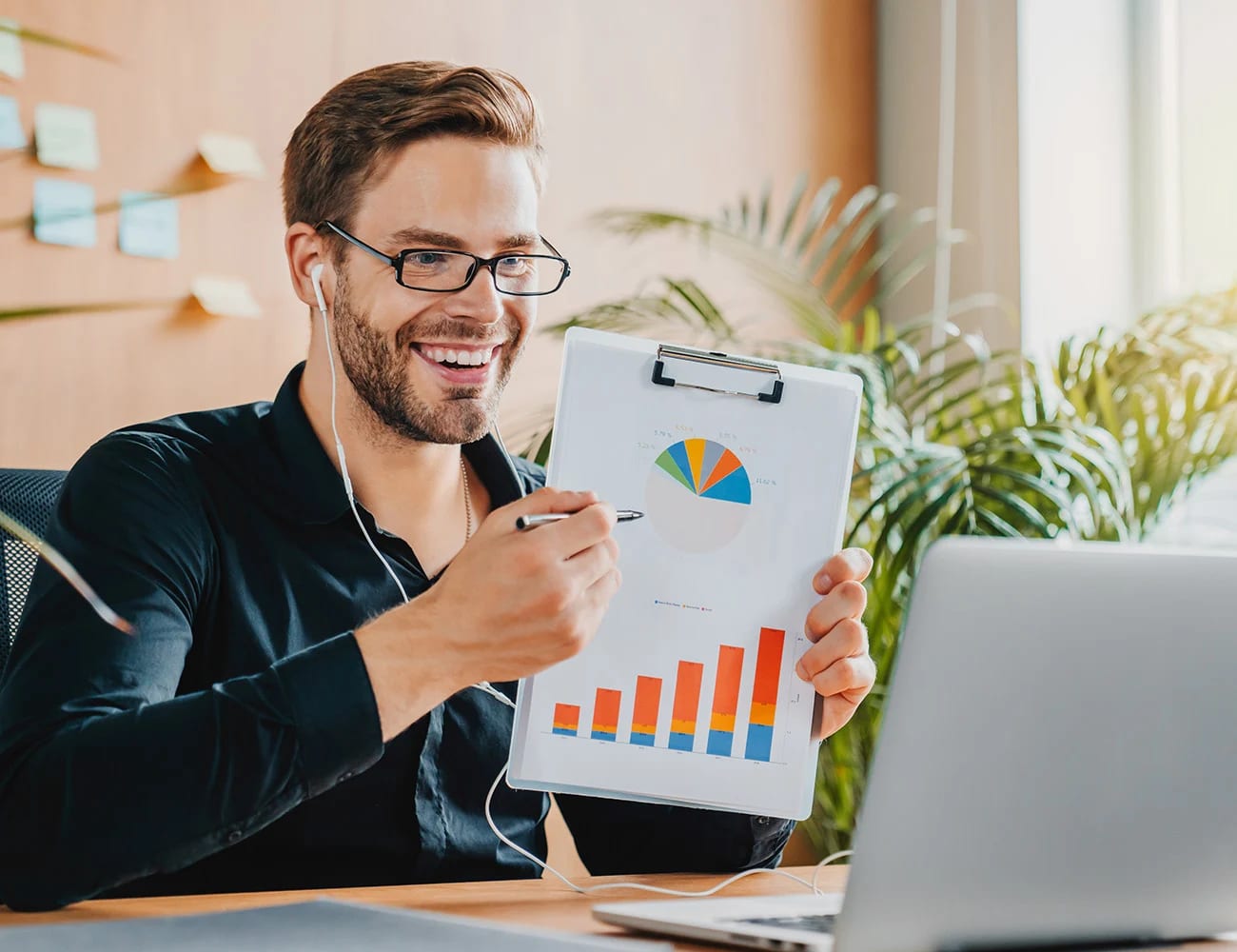 Person at a desk holding up a clipboard with a bar chart and a pie chart beside a laptop